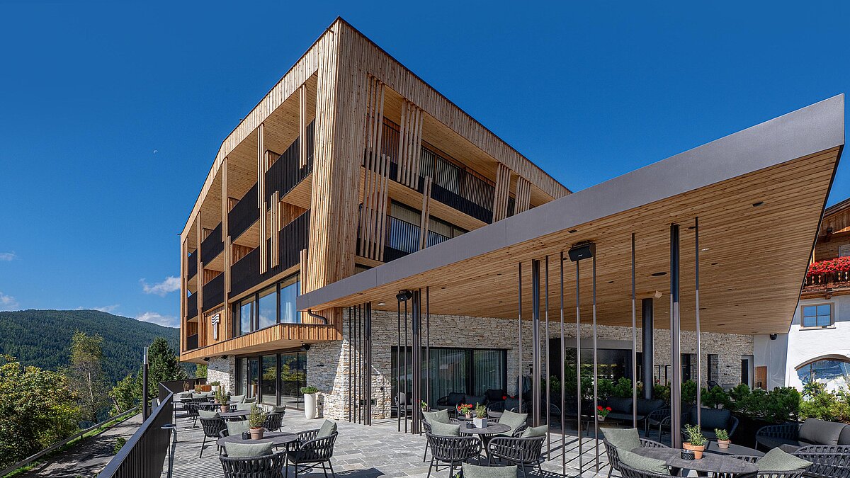 Modernes Hotelgebäude aus Holz und Stein mit weitläufiger Terrasse, bestückt mit runden Tischen und Stühlen. Umgeben von üppiger Natur und blauen Himmel, bietet die Terrasse entspannte Sitzgelegenheiten mit Pflanzen.