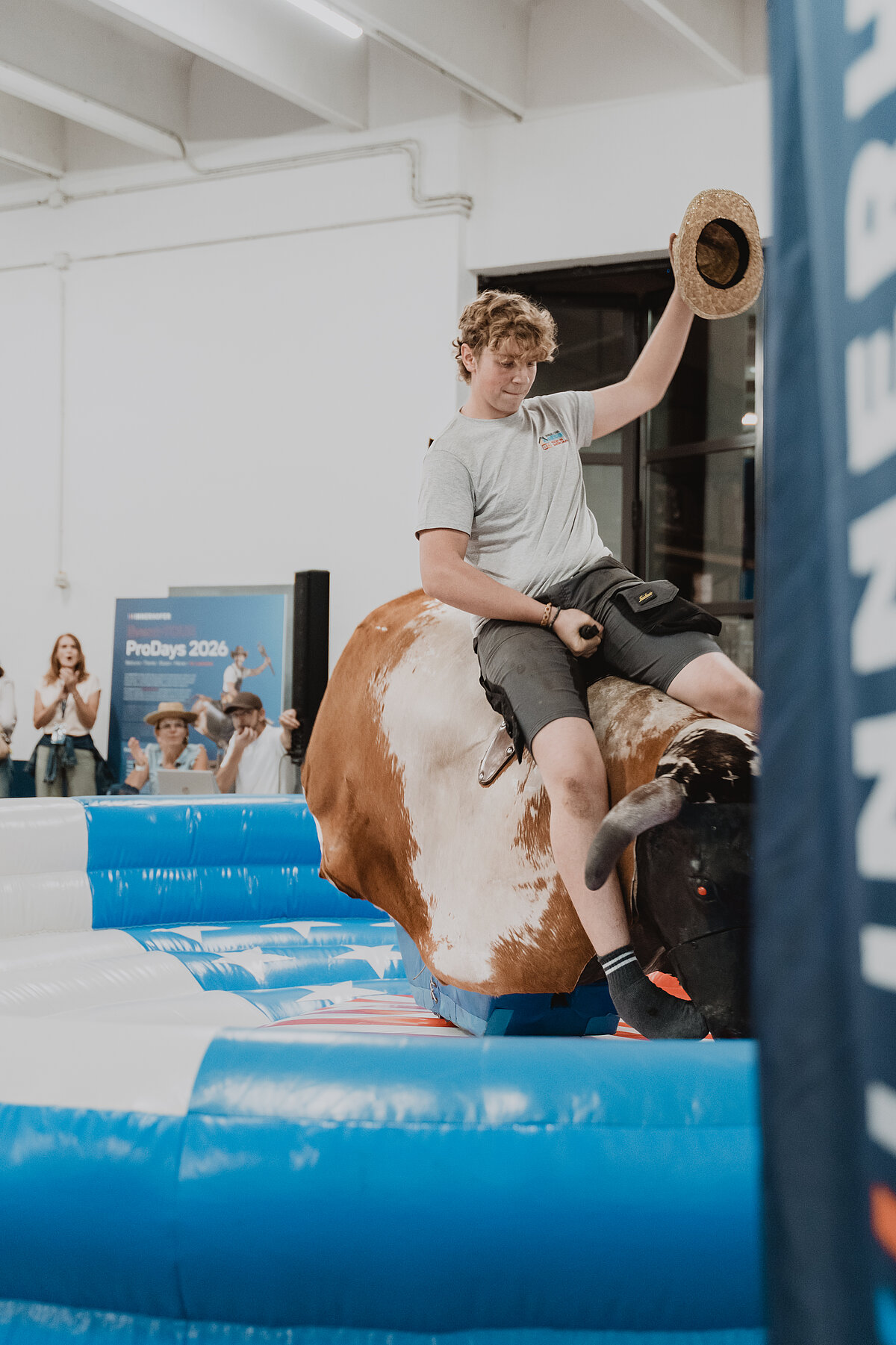 Ein Mann reitet auf einem mechanischen Bullen in einer Halle und hält dabei einen Hut in der Hand. Um ihn herum schauen mehrere Personen zu. Der Bullenreitplatz besteht aus einem blau-weißen aufblasbaren Kissen.