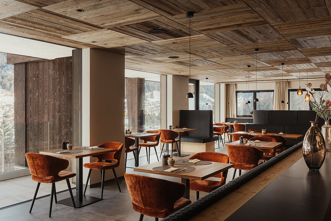 Ein modernes Restaurant mit Holzelementen, großen Fenstern und warmen braunen und orangefarbenen Stühlen. Tische sind sauber gedeckt, und die Beleuchtung ist gedämpft. Ein eleganter, ruhiger Essbereich mit dekorativen Blumenvasen.