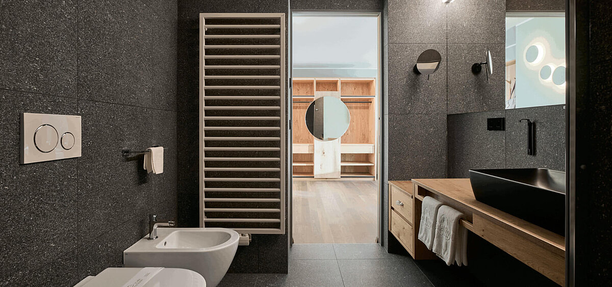 Modernes Badezimmer mit dunkler Steinwand, großem Spiegel, Holzschränken und Badewanne. Im Hintergrund ist ein Flur mit Tageslicht zu sehen.