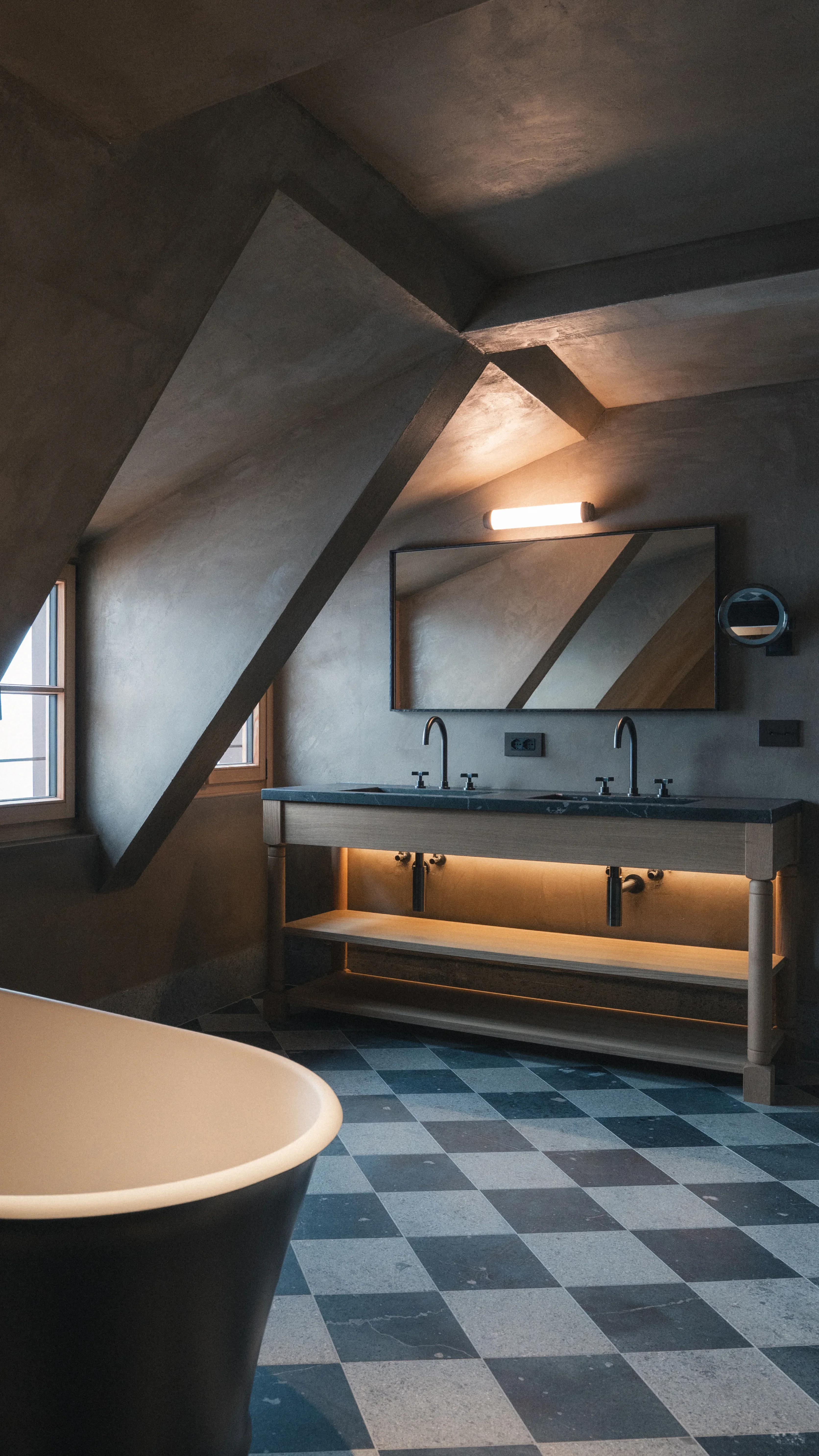 Modernes Badezimmer mit grauem und schwarzem Schachbrettboden, Doppelwaschbecken aus Stein, großem rechteckigem Spiegel und Wandleuchte darüber. Eine freistehende Badewanne steht im Vordergrund, Dachschrägen säumen den Raum.