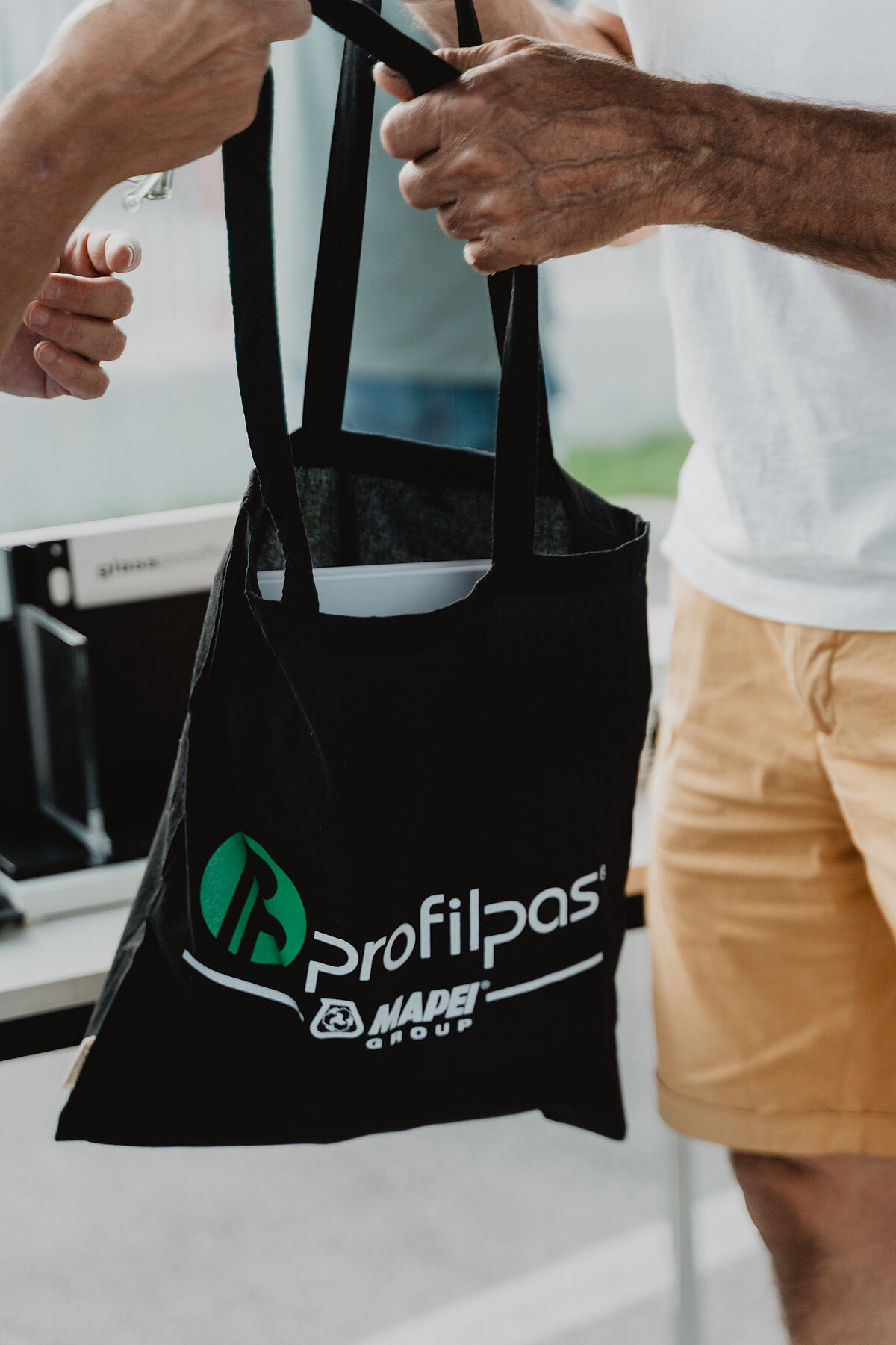 Zwei Personen tauschen eine schwarze Stofftasche mit dem Aufdruck "Profilpas" aus. Eine Person trägt ein weißes T-Shirt und gelbe Shorts. Die Szene findet im Freien statt.