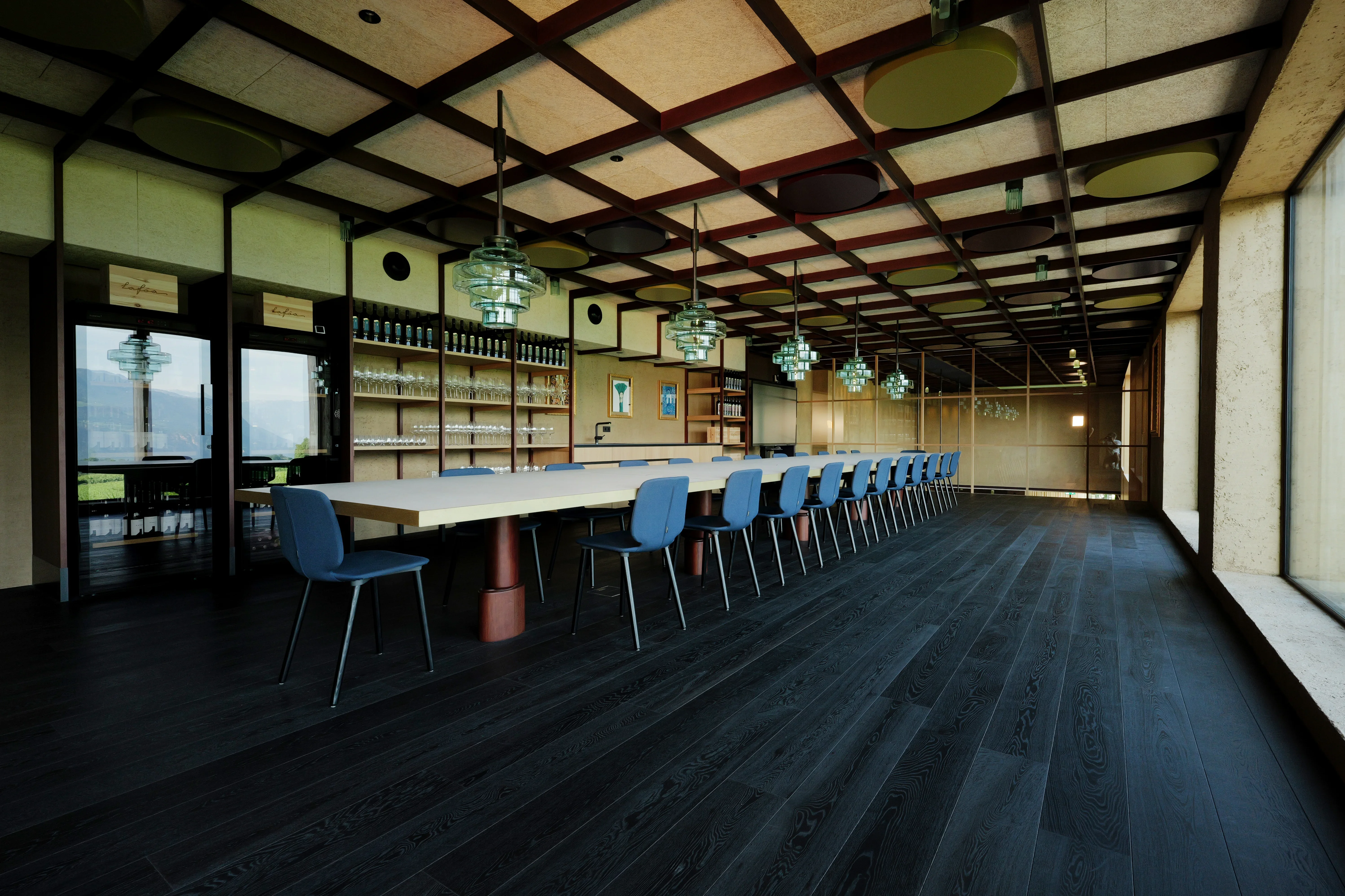 Ein großer, moderner Konferenzraum mit einem langen Tisch und blauen Stühlen. Der Raum hat eine Decke mit Holzgitter, Hängelampen und große Fenster, die viel Licht hereinlassen. An der Rückwand befinden sich Regale mit Flaschen und andere Dekorationselemente.