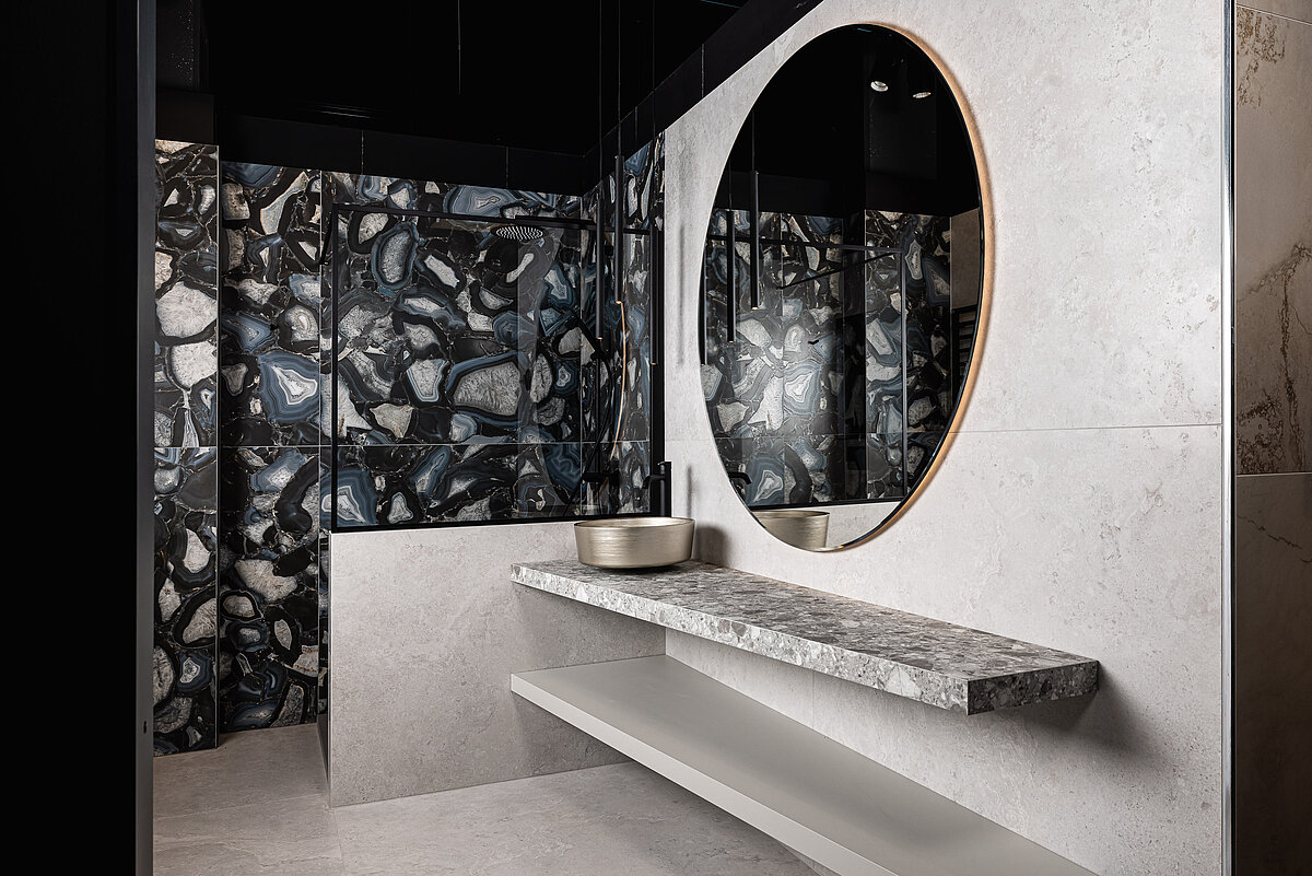 Modernes Badezimmer mit großen Marmorfliesen, einem runden Spiegel und einem modernen Steinwaschbecken auf einem eleganten, grauen Waschtisch. Die Wände sind mit schwarzen und weißen Mosaikfliesen gestaltet. Minimalistische Einrichtung mit Fokus auf natürliche Materialien und klare Linien.