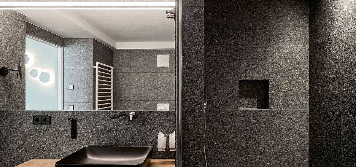 Modernes Badezimmer mit dunkler Steinwand, großem Spiegel, Holzschränken und Badewanne. Im Hintergrund ist ein Flur mit Tageslicht zu sehen.