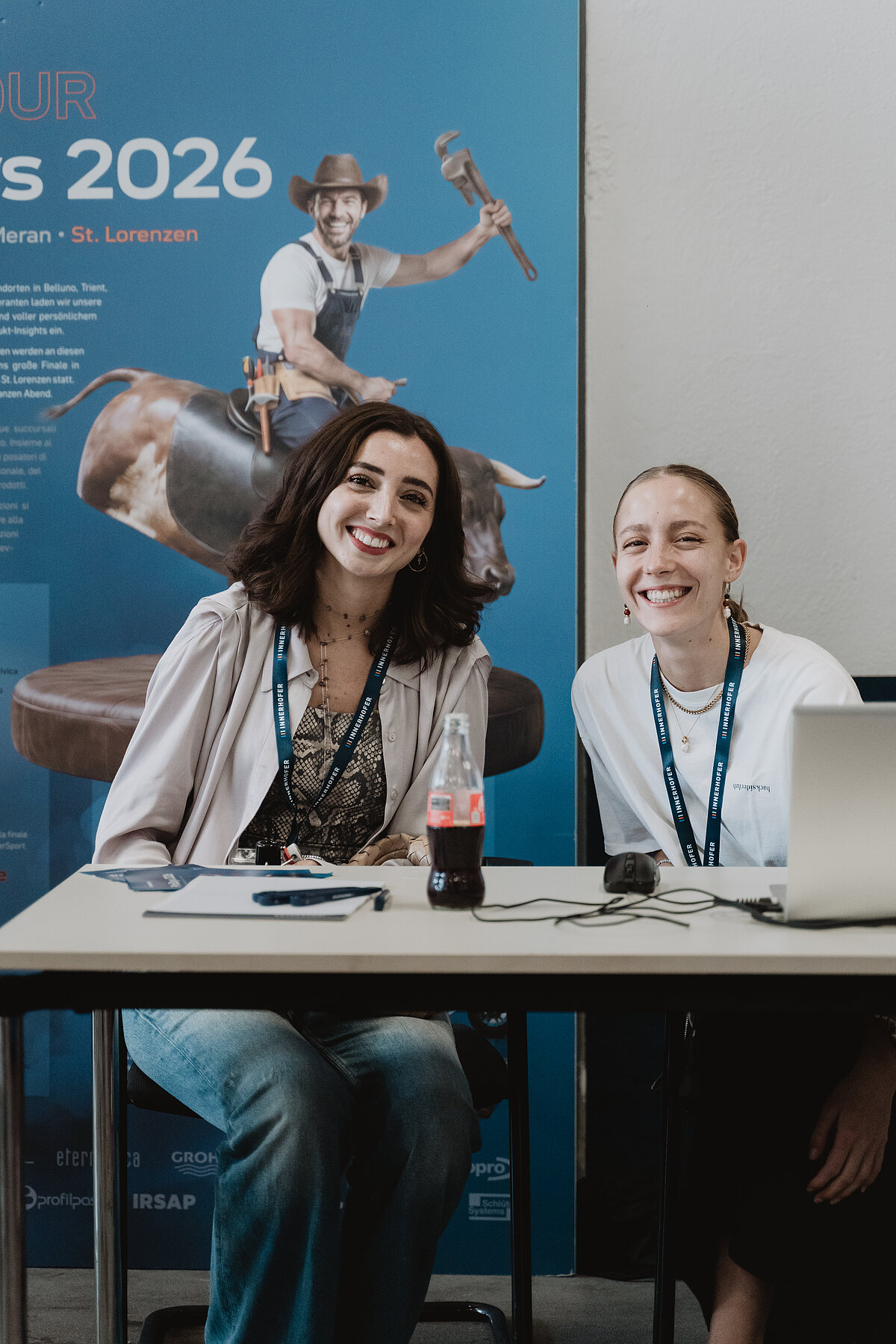 Zwei lachende Frauen sitzen an einem Tisch mit einem Laptop und einer Colaflasche. Im Hintergrund ist ein Plakat mit einem Mann auf einem mechanischen Bullen zu sehen, das Werbung für eine Veranstaltung im Jahr 2026 macht.