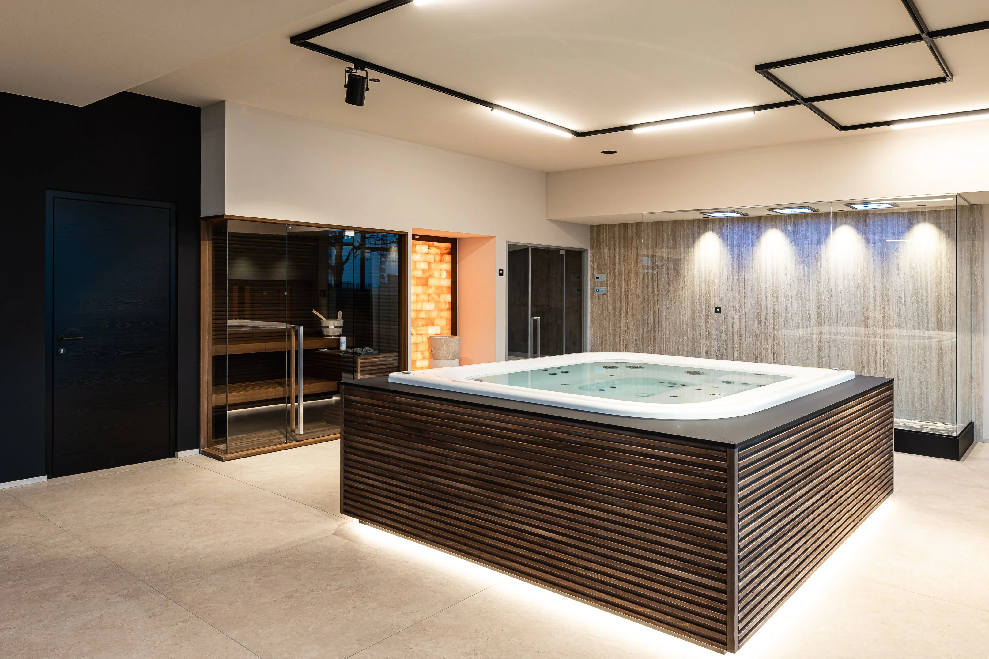 Luxuriöser Wellnessbereich mit großem Whirlpool, moderner Holzverkleidung und schwarz gefliester Wand. Im Hintergrund sind eine Sauna mit Glasfront und ein Duschbereich mit beleuchteten Fliesen zu sehen. Dezente Beleuchtung verleiht dem Raum eine entspannende Atmosphäre.