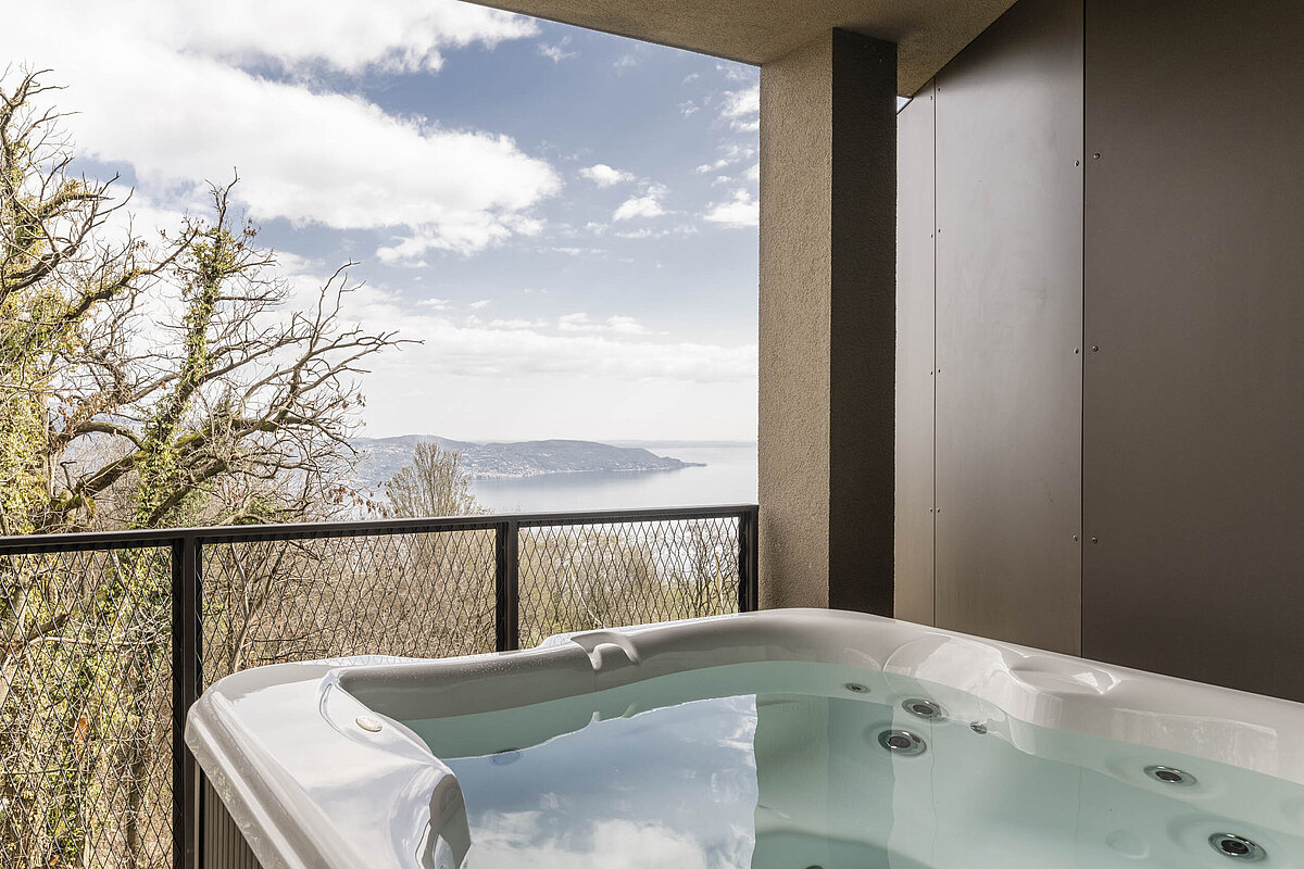 Ein Whirlpool auf einem Balkon bietet einen Ausblick auf einen bewaldeten Hügel und einen ruhigen See unter einem bewölkten Himmel.