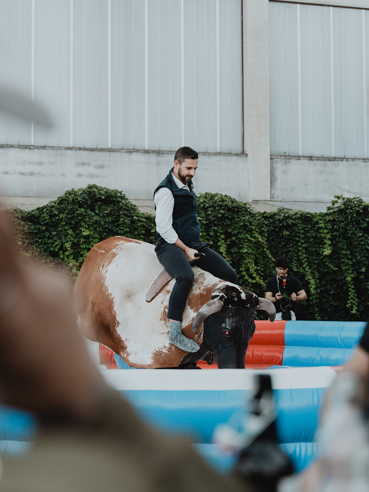 Ein Mann reitet auf einem mechanischen Bullen in einem aufblasbaren Ring im Freien. Um ihn herum stehen Zuschauer und im Hintergrund ist eine mit Efeu bewachsene Mauer zu sehen.
