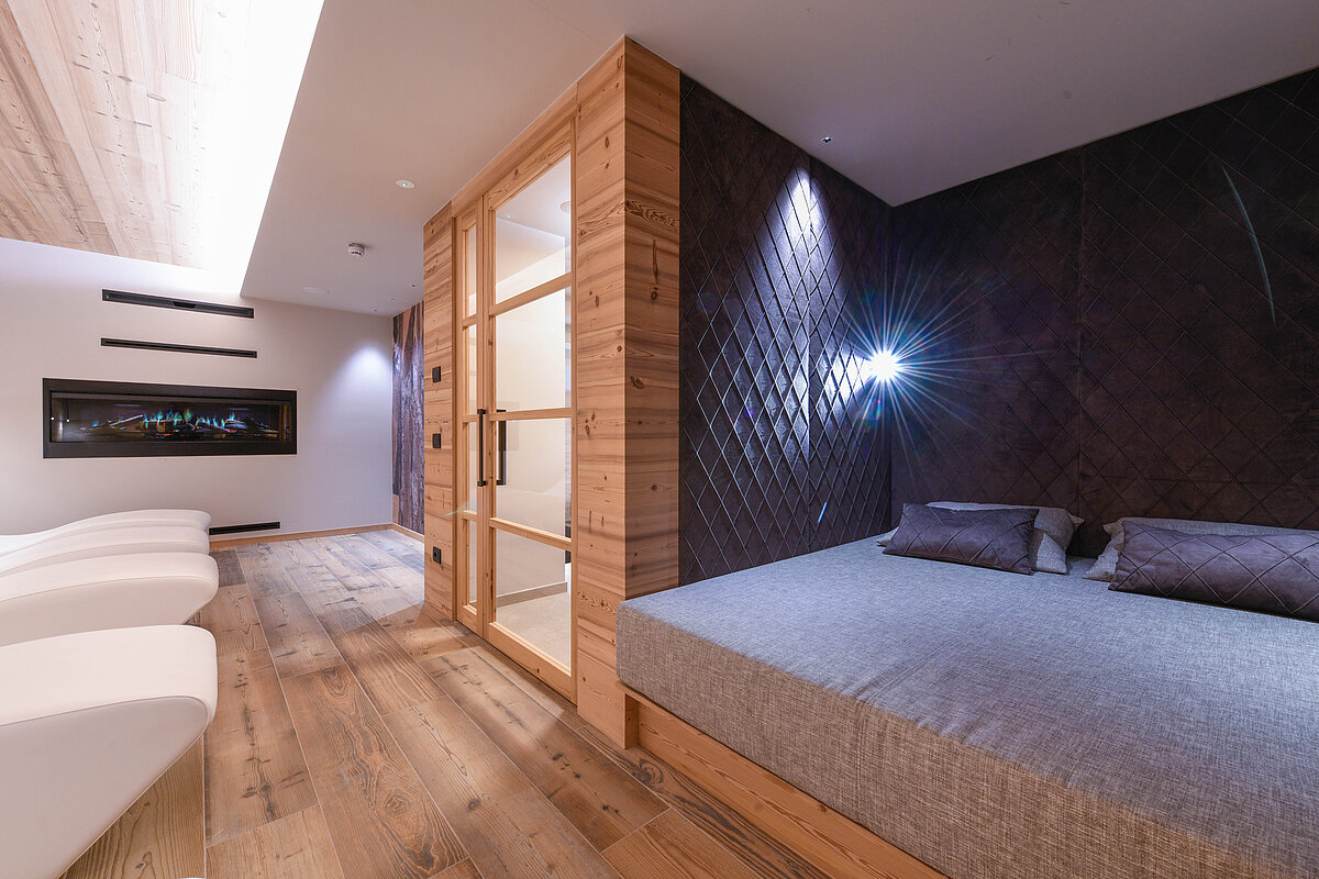 Moderner Wellnessbereich mit Holzelementen, bestehend aus einer Glas-Saunakabine, großen grauen Liegen und einer indirekt beleuchteten Holzwand. Der Raum hat einen Holzboden und stilvolle Beleuchtung, die eine entspannende Atmosphäre schafft.