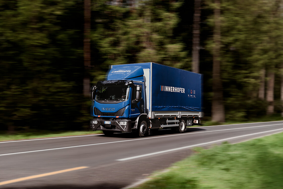 Ein blauer LKW mit der Aufschrift "INNERHOFER" fährt auf einer Landstraße, umgeben von Bäumen. Der LKW ist in Bewegung und die Straße verläuft durch eine grüne, bewaldete Landschaft.