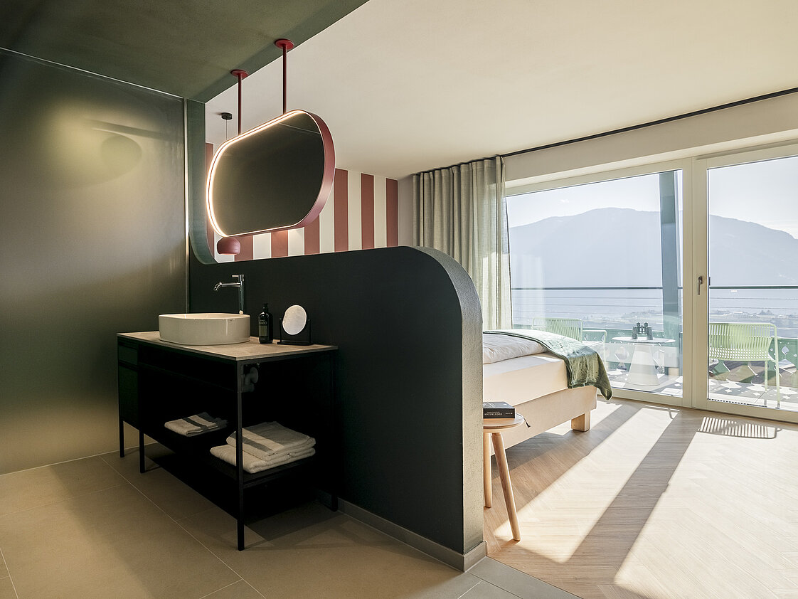 Modernes Hotelzimmer mit offenem Badezimmerbereich. Ein Waschbecken mit einem großen beleuchteten Spiegel ist am linken Rand, während ein Bett mit grüner Decke und Aussicht auf Berge durch ein großes Fenster zu sehen ist. Das Zimmer wirkt hell und minimalistisch.