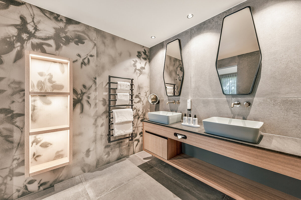 Modernes Badezimmer mit zwei rechteckigen Waschbecken auf einer hölzernen Waschtischplatte, zwei markanten Spiegeln an der Wand und einem dekorativen Handtuchhalter. Die Wände sind mit einem floralen Muster verziert. Dezente Beleuchtung sorgt für eine entspannte Atmosphäre.
