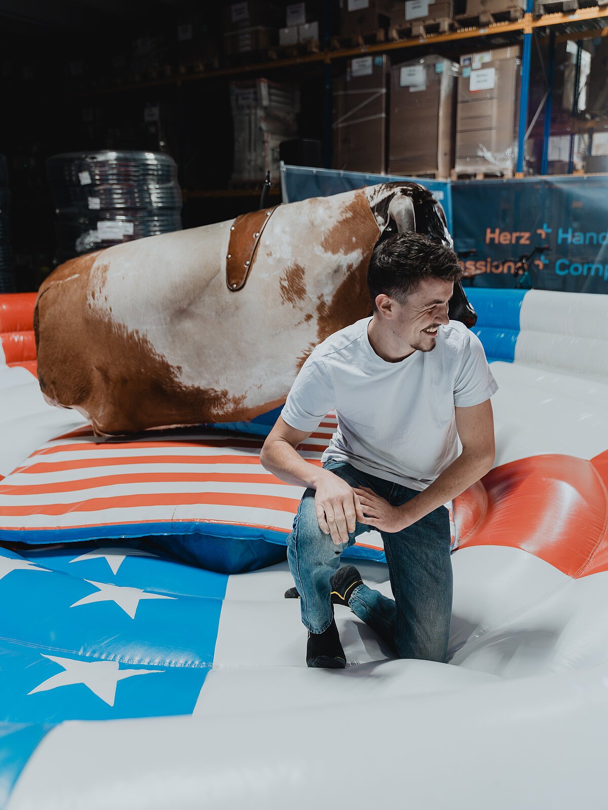 Ein Mann in einem weißen T-Shirt und Jeans kniet lachend auf einem mechanischen Bullen, umgeben von einer aufblasbaren Matte im USA-Flaggen-Design. Im Hintergrund sind Kartons und Regale zu sehen.
