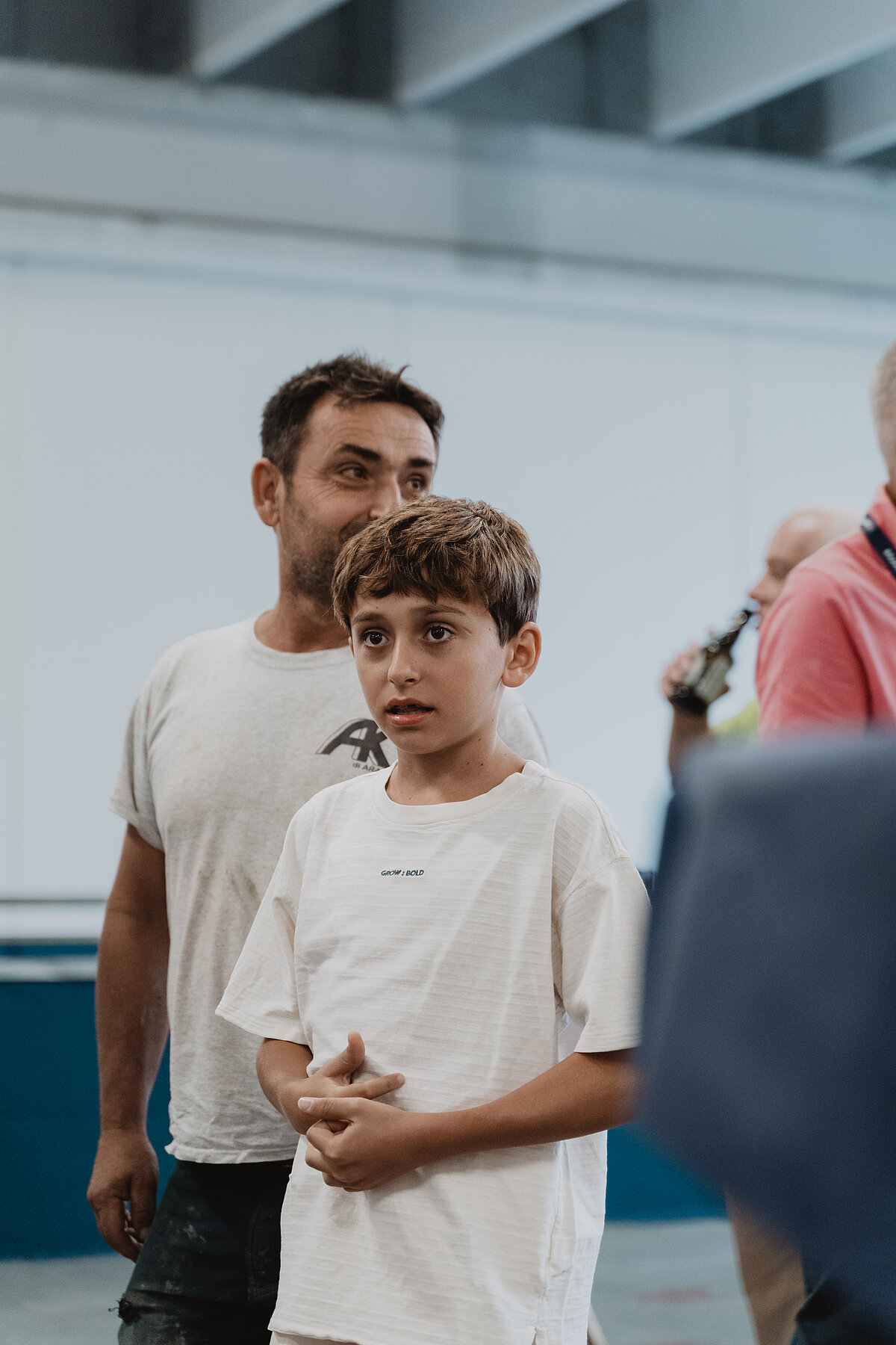 Ein Junge und ein Mann stehen zusammen in einem Innenraum mit neutralem Hintergrund. Der Junge trägt ein weißes T-Shirt und schaut interessiert nach vorne. Der Mann steht hinter ihm und trägt ebenfalls ein helles T-Shirt. Im Hintergrund ist eine weitere Person teilweise sichtbar.