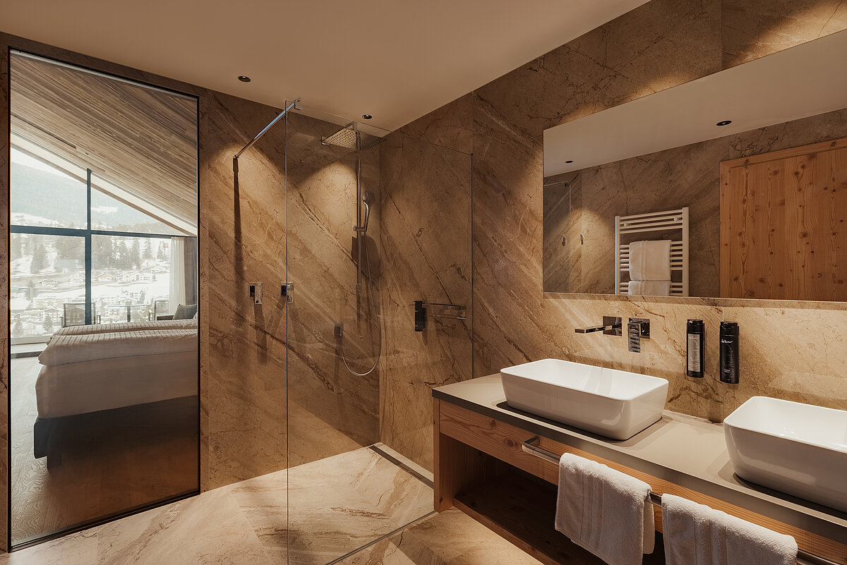 Modernes Badezimmer mit zwei weißen Waschbecken auf einem Holzunterschrank, großem Spiegel, Glasdusche und Fenster mit Blick auf ein Bett und die Außenlandschaft. Helle, marmorne Wände und Böden verleihen dem Raum ein luxuriöses Ambiente.