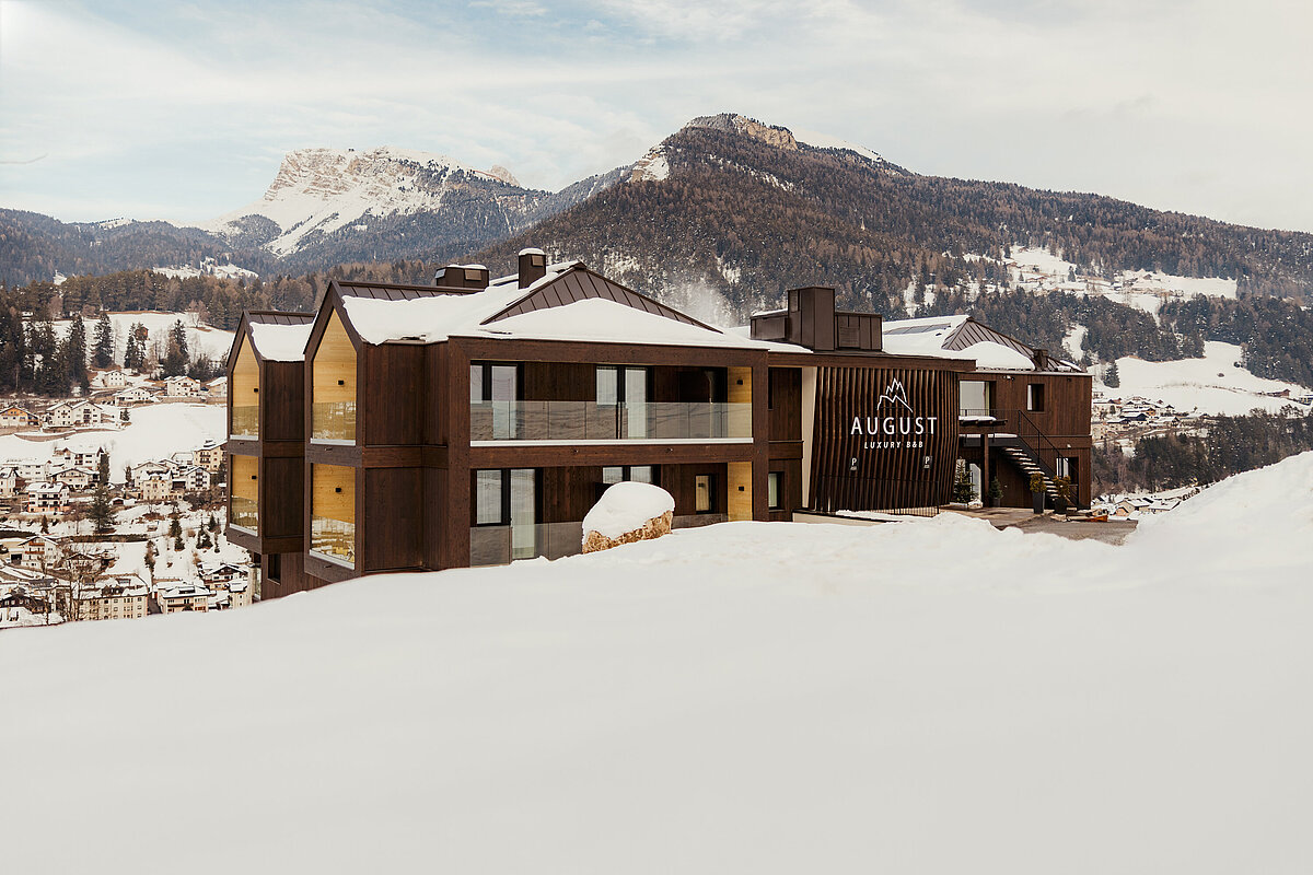 Ein modernes Hotelgebäude mit dunkler Fassade, gelegen in einer verschneiten Berglandschaft. Im Hintergrund sind schneebedeckte Berge und ein Tal mit einem Dorf sichtbar. Auf dem Gebäude steht „AUGUST“ geschrieben.