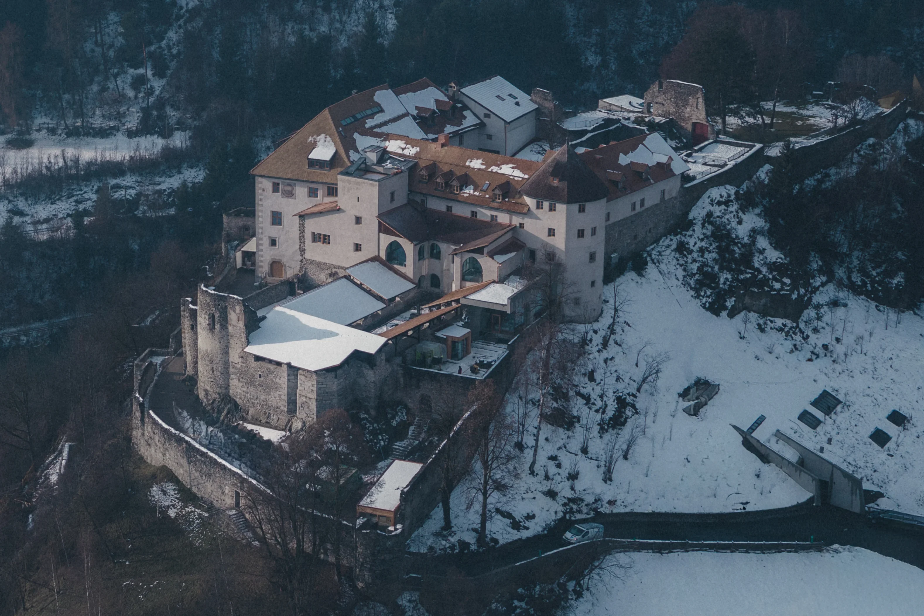 Luftaufnahme eines alten Schlosses auf einem Hügel inmitten eines schneebedeckten Waldes. Die Gebäude sind von dicken Steinmauern umgeben. Teile des Dachs sind mit Schnee bedeckt. Im Vordergrund ist ein gefrorener und schneebedeckter Fluss oder See sichtbar.