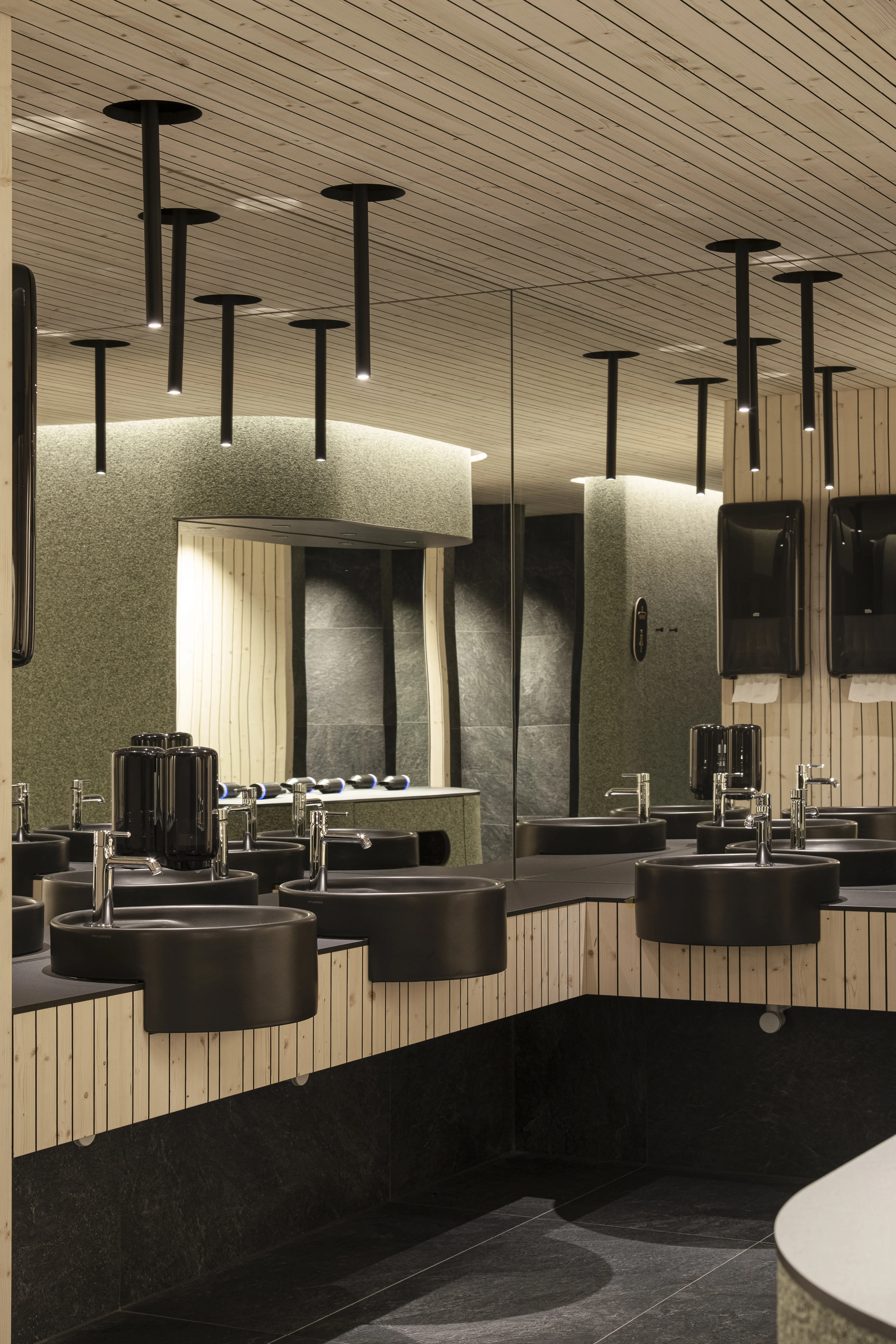 Moderner Waschraum mit stilvollem Design. Schwarze runde Waschbecken mit eleganten Armaturen auf glatten Oberflächen. Hängende, zylinderförmige Leuchten an der Holzdecke. Große Spiegel hinter den Waschbecken reflektieren den Raum. Dezente Beleuchtung und glatte Wände schaffen eine entspannte Atmosphäre.