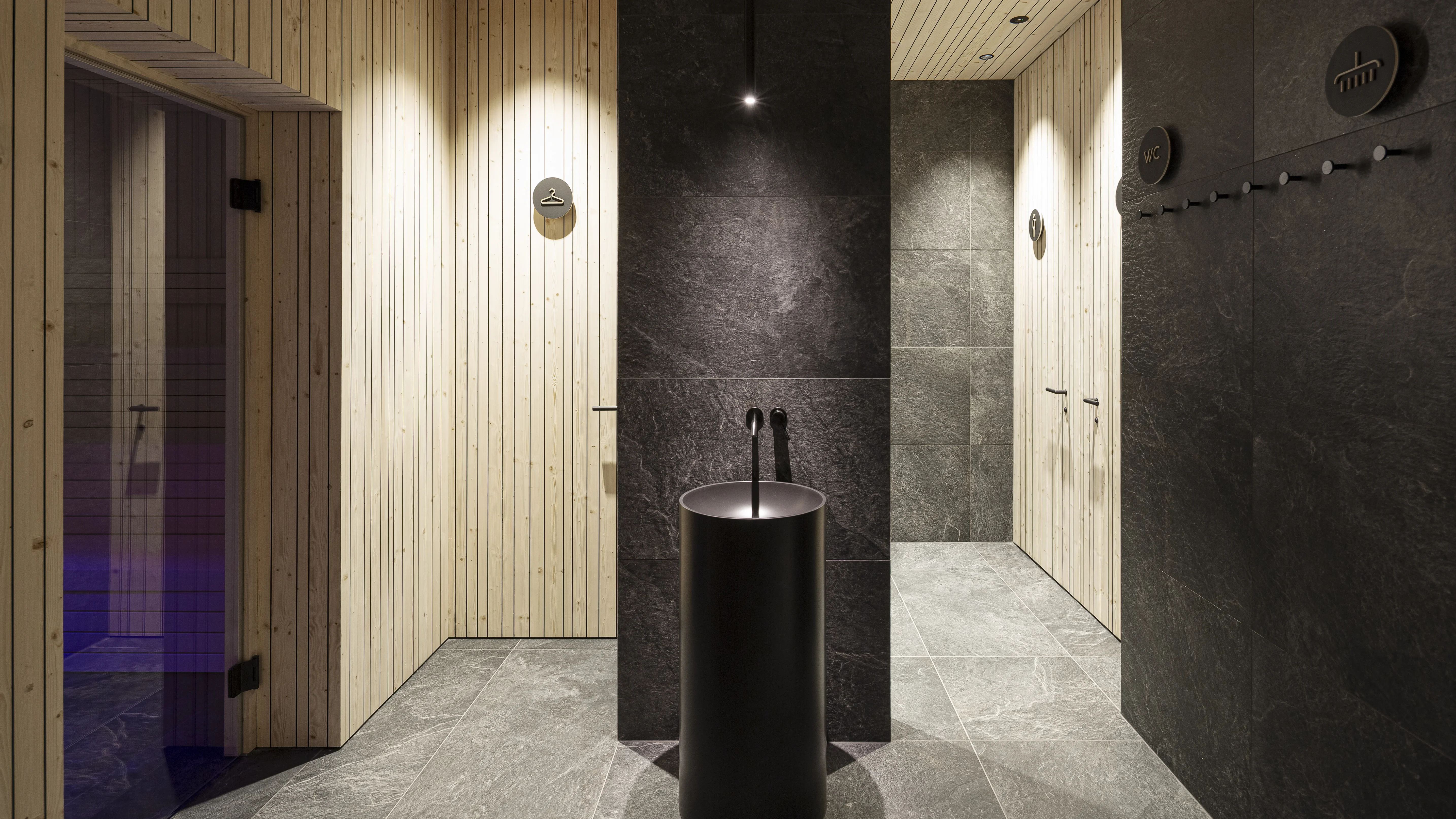 Moderner Saunavorraum mit dunklem Bodenbelag und Wänden. Ein schwarzes, zylindrisches Waschbecken steht im Zentrum vor einer dunklen Wand. Links führt eine Tür in einen erleuchteten Saunabereich. Holzverkleidete Wände sorgen für einen warmen Kontrast.