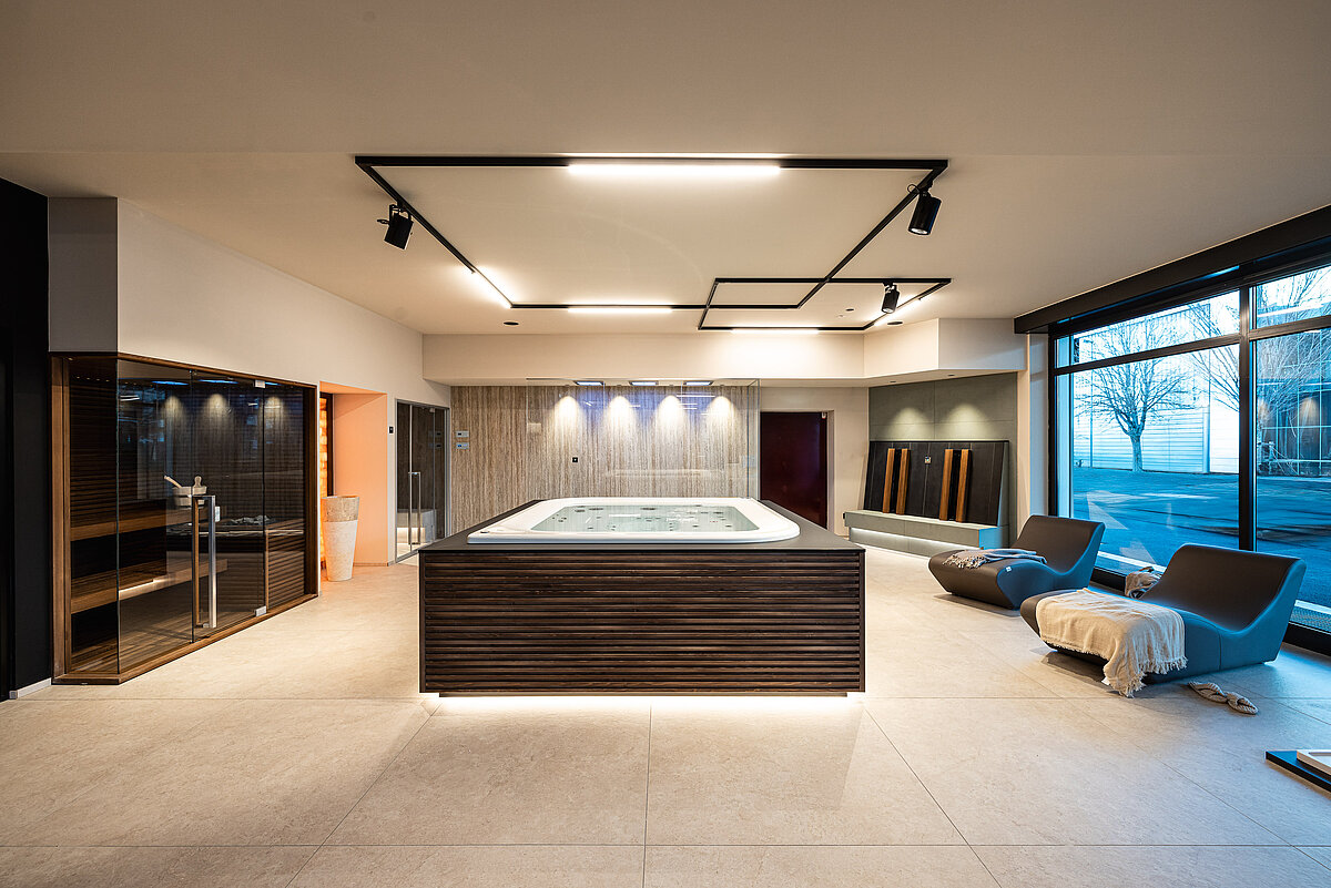 Ein moderner Wellnessbereich mit einem zentralen großen Whirlpool, eleganten Loungesesseln mit Decke, einer Glasdusche und gedämpfter Beleuchtung. Große Fenster bieten einen Ausblick nach draußen. Der Raum strahlt eine entspannende und luxuriöse Atmosphäre aus.