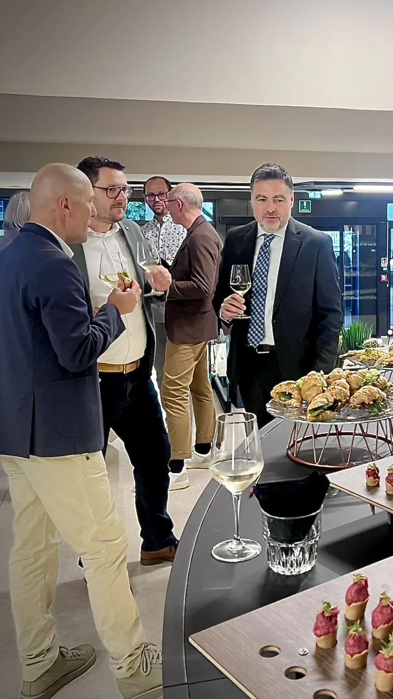 Mehrere Männer in eleganter Kleidung stehen an einem Buffet mit verschiedenen Appetithäppchen und unterhalten sich. Einige halten Weingläser in der Hand. Die Atmosphäre wirkt gehoben und entspannt.