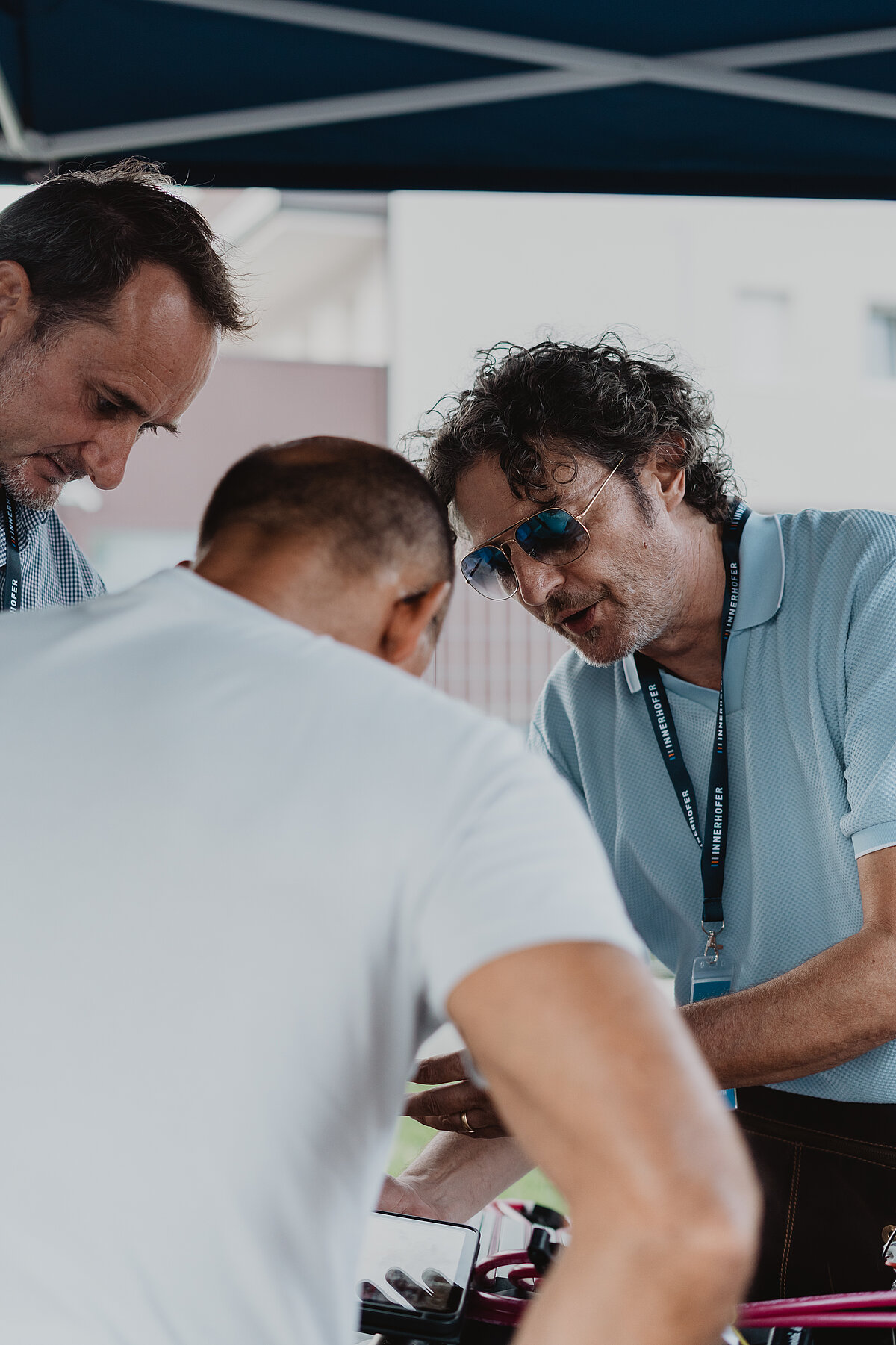 Drei Männer stehen unter einem blauen Zelt und unterhalten sich. Der Mann in der Mitte trägt eine Sonnenbrille und ein Namensschild um den Hals. Die Männer scheinen konzentriert und engagiert in ihre Diskussion vertieft zu sein.