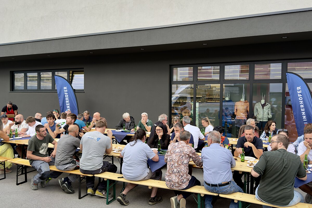 Menschen sitzen an langen Bänken draußen vor einem modernen Gebäude und essen. Es herrscht eine gesellige und entspannte Atmosphäre. Im Hintergrund sind Fahnen mit dem Schriftzug „Zahnen“ zu sehen.