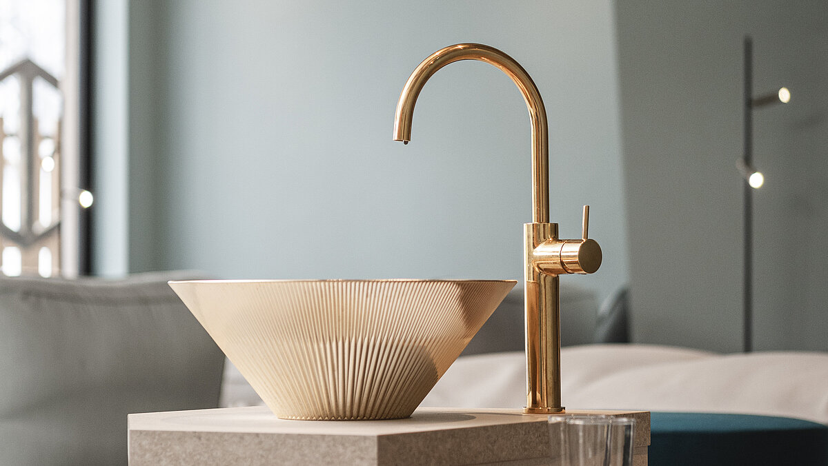 Ein modernes Badezimmer mit einem eleganten, goldenen Wasserhahn und einem stilvollen Waschbecken aus porzellanartigem Material. Auf der Holzfläche neben dem Becken steht ein durchsichtiges Glas. Die Wände sind hellblau gehalten und im Hintergrund ist ein Fenster zu sehen.