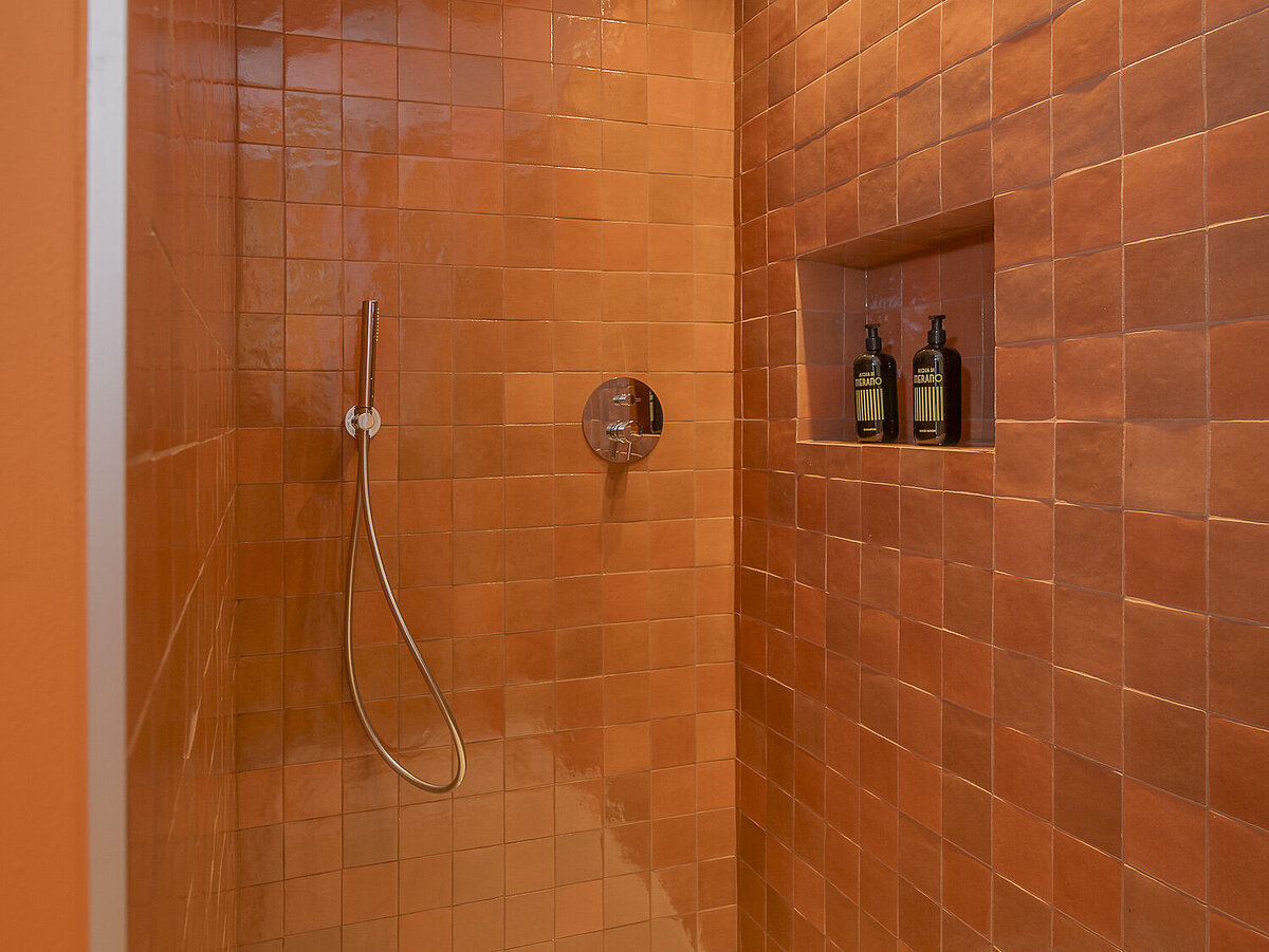 Eine Dusche mit orangefarbenen Fliesen, einer Regendusche und einem Handbrausekopf. In einer eingebauten Nische stehen Pflegeprodukte. Ein minimalistisches und modernes Design.