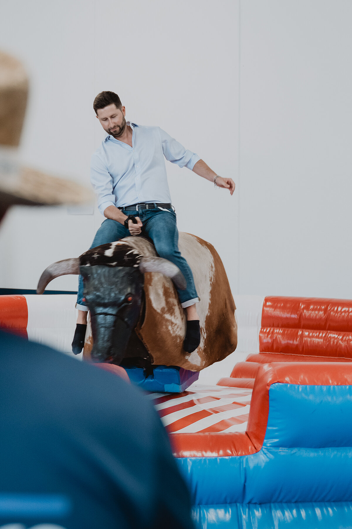 Ein Mann reitet auf einem mechanischen Bullen in einer indoor Umgebung. Er trägt ein hellblaues Hemd und Jeans. Im Vordergrund ist unscharf ein Zuschauer mit einem Hut zu sehen. Die Umgebung ist mit bunten Polstern ausgestattet.
