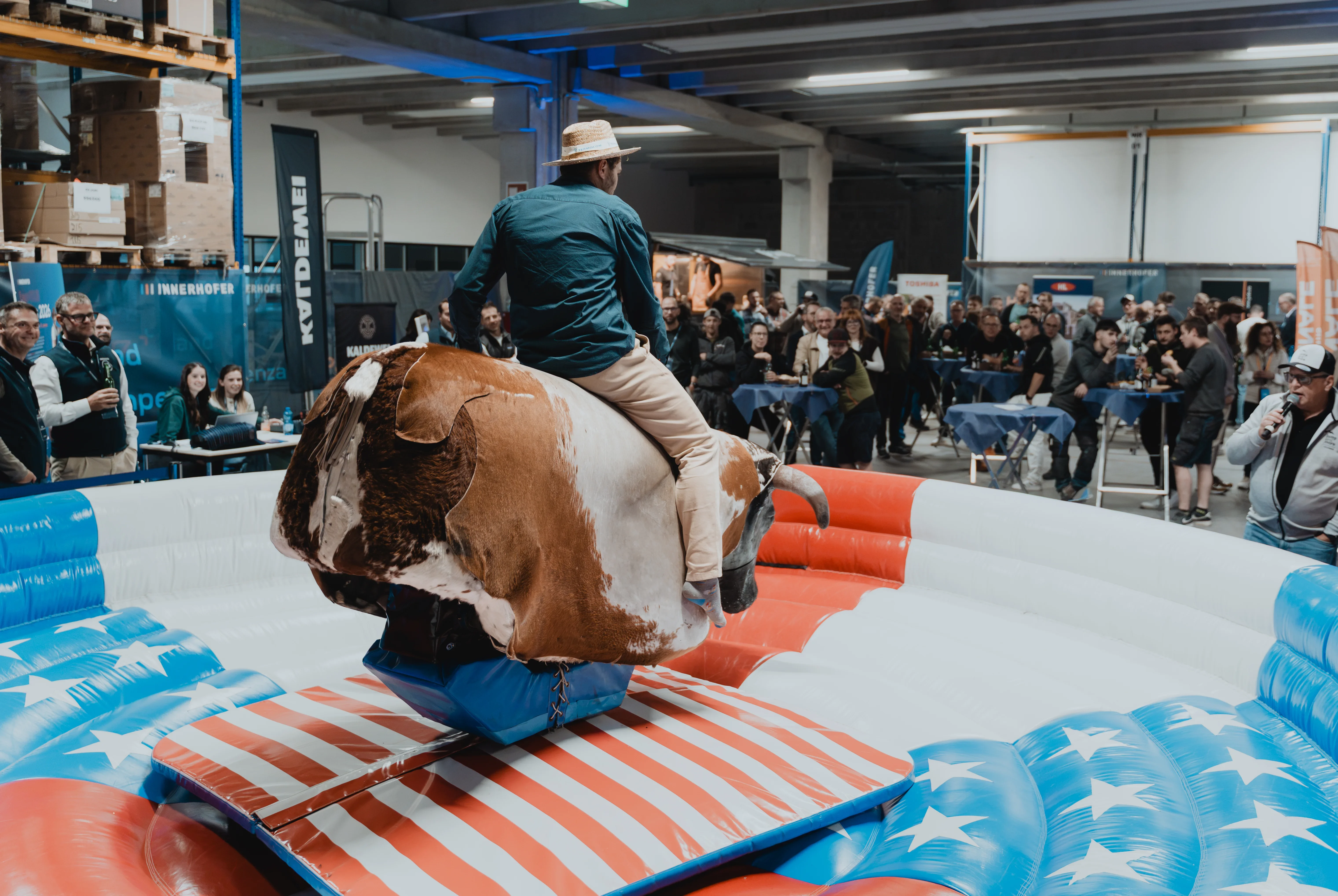 Eine Person reitet auf einem mechanischen Bullen in einer Halle. Um den Bullen herum ist eine gepolsterte Struktur mit amerikanischem Flaggenmotiv. Im Hintergrund stehen zahlreiche Zuschauer, die dem Geschehen zusehen.