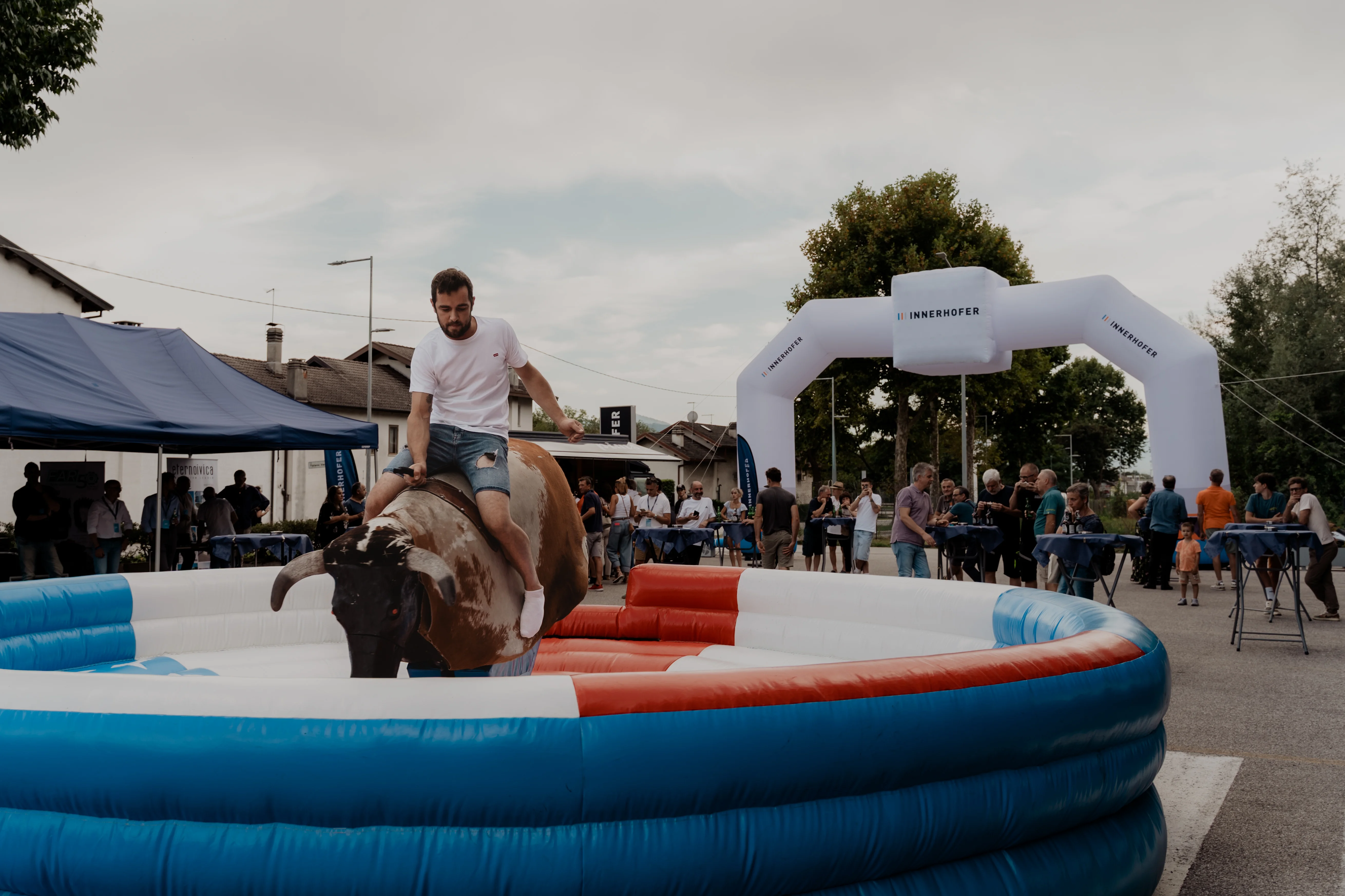 Ein Mann reitet auf einem mechanischen Bullen in einer aufblasbaren Arena bei einer Outdoor-Veranstaltung. Im Hintergrund stehen Zelte und eine Gruppe von Menschen, die das Geschehen beobachten. Es sind Bäume und ein Bogen mit der Aufschrift "BRENNERSTAHL" sichtbar.
