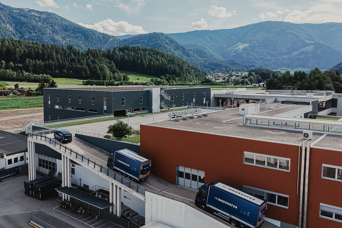 Luftaufnahme eines großen Fabrikgeländes mit verschiedenen Industriegebäuden in einer bergigen Landschaft. Mehrere Liefer-Lastwagen sind auf einer Rampe zu sehen. Im Hintergrund stehen bewaldete Hügel und ein wolkiger Himmel.