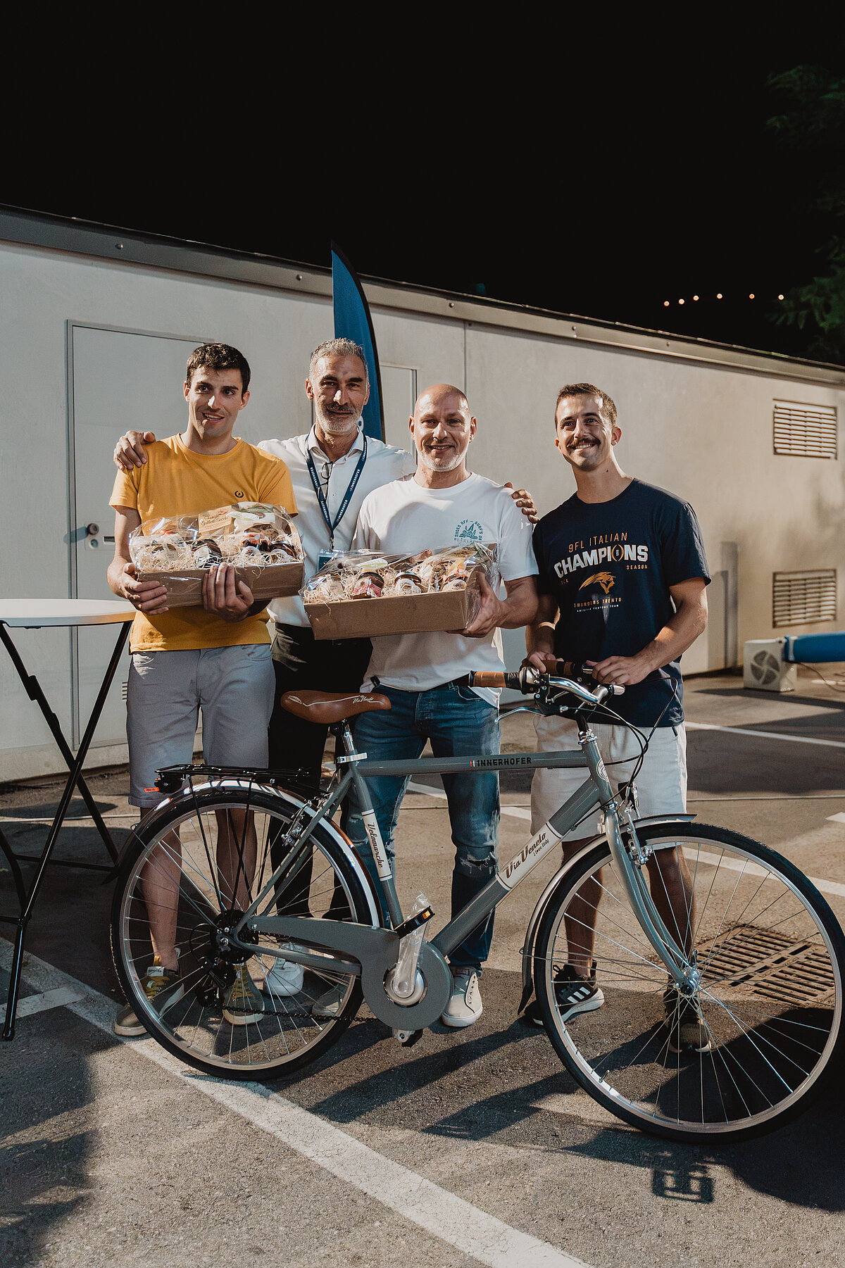 Vier Männer posieren draußen mit einem Fahrrad. Zwei von ihnen halten Tabletts mit Backwaren. Sie stehen vor einem Gebäude, es ist Nacht. Alle lächeln in die Kamera.
