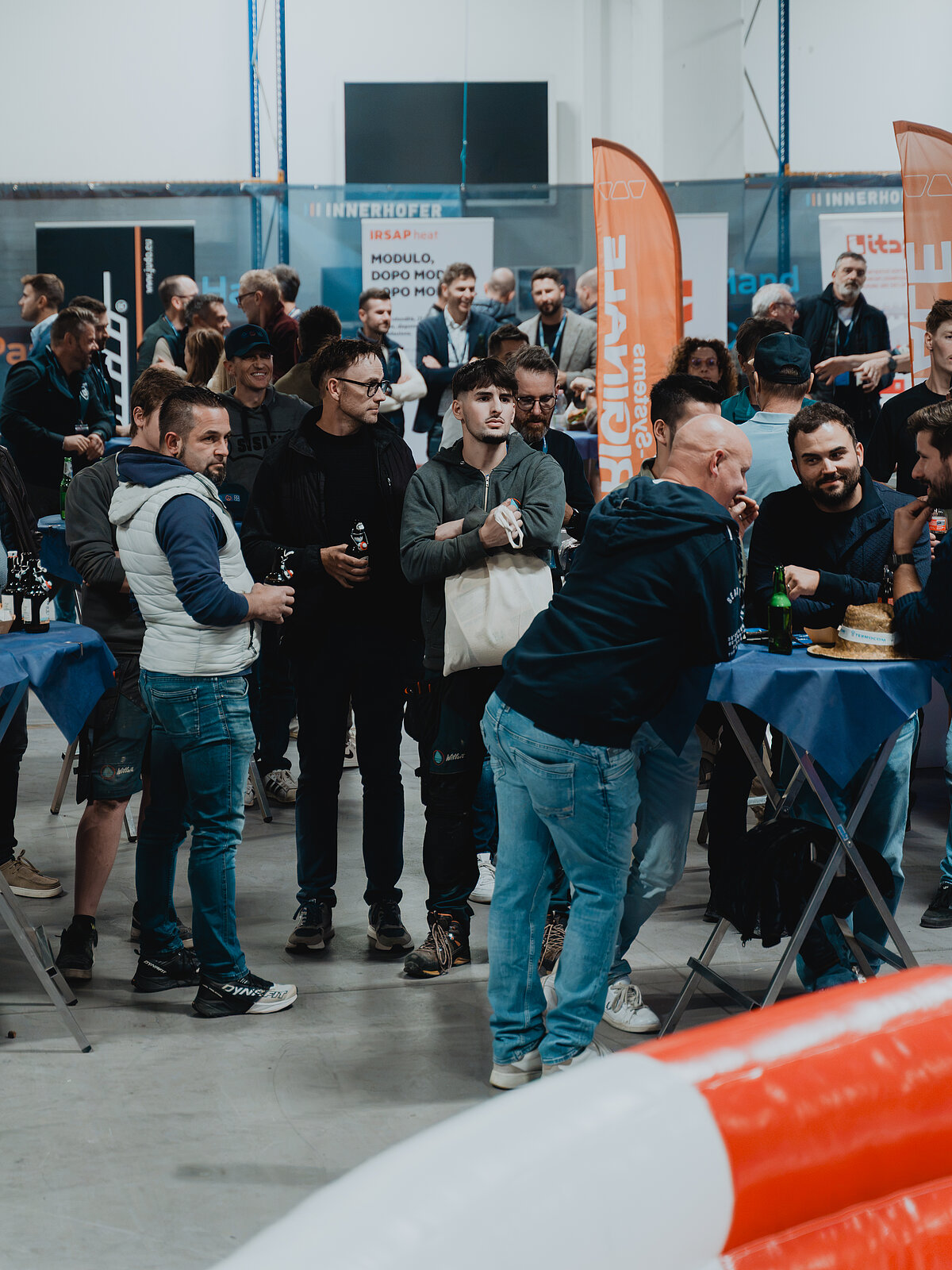 Mehrere Personen stehen in einer Halle um Tische herum. Sie scheinen an einer informellen Veranstaltung teilzunehmen, einige halten Getränke in der Hand. Im Hintergrund sind Banner und Stände zu sehen. Die Atmosphäre ist lebhaft und gesellig.