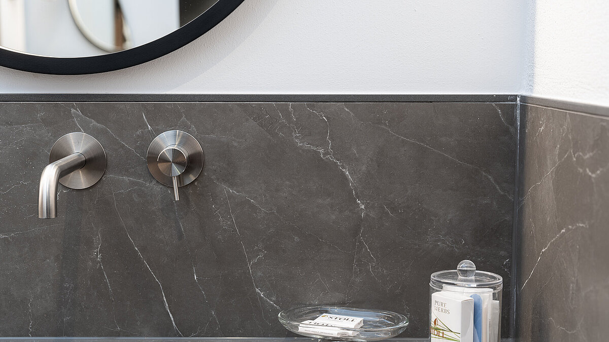 Moderner Badezimmerschrank mit grauer Steinoberfläche. An der Wand ein minimalistischer Wasserhahn und ein Knopf aus Edelstahl. Auf dem Schrank stehen zwei Gläser: eins mit Seife und eins mit Zahnbürsten. Ein gefaltetes Handtuch liegt daneben.