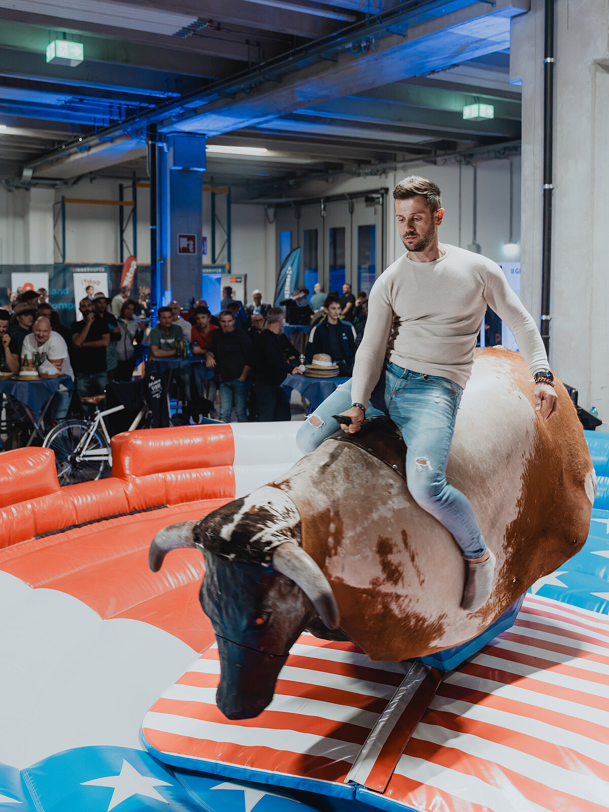 Ein Mann reitet auf einem mechanischen Bullen in einer Halle, umgeben von einer Sicherheitsmatte mit amerikanischem Flaggenmuster. Im Hintergrund beobachtet eine Gruppe von Menschen das Geschehen.