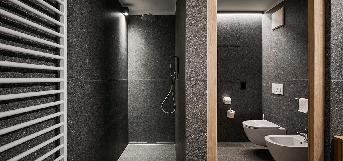 Modernes Badezimmer mit dunkler Steinwand, großem Spiegel, Holzschränken und Badewanne. Im Hintergrund ist ein Flur mit Tageslicht zu sehen.