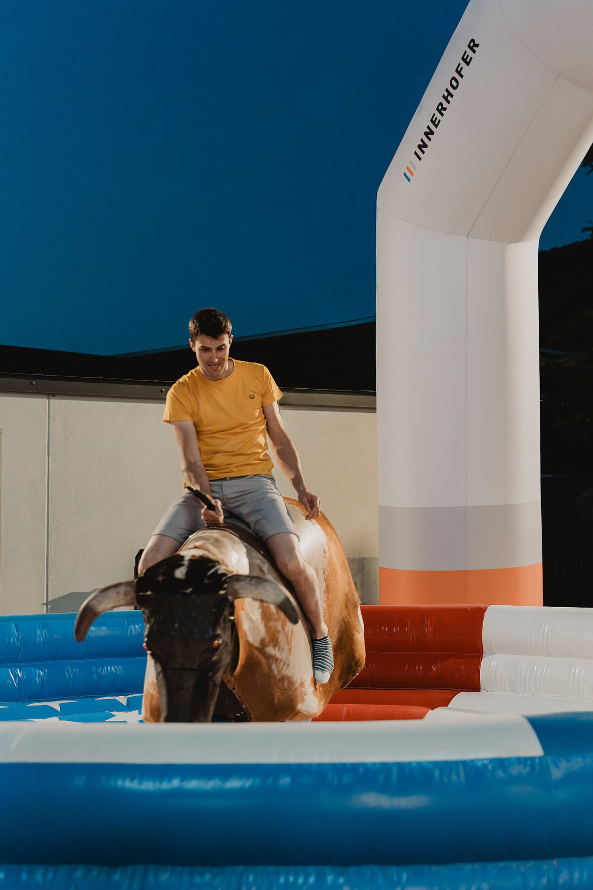 Ein Mann in einem gelben T-Shirt reitet auf einem mechanischen Bullen in einer aufblasbaren Arena. Es ist Abend, und das Licht ist gedämpft. Im Hintergrund ist ein Werbebanner von „Innerhofer“ zu sehen.