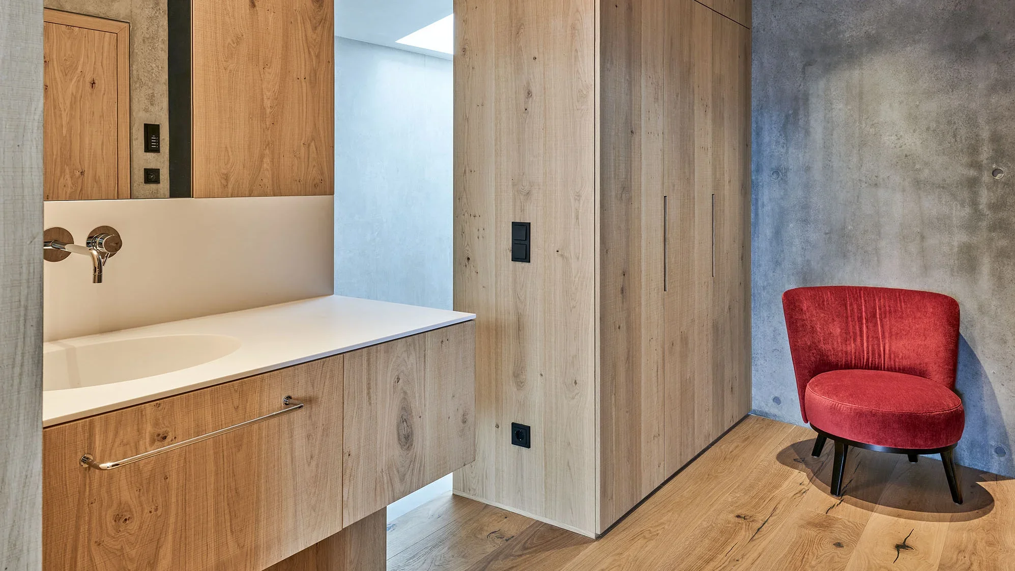 Modernes Badezimmer mit Holzverkleidung, einer weißen Waschtischplatte mit integriertem Wasserhahn, und einem roten Stuhl in der Ecke. Das Zimmer wirkt hell und minimalistisch mit einer warmen Holzoptik auf Boden und Schränken. Wände in Betonoptik ergänzen das Design.