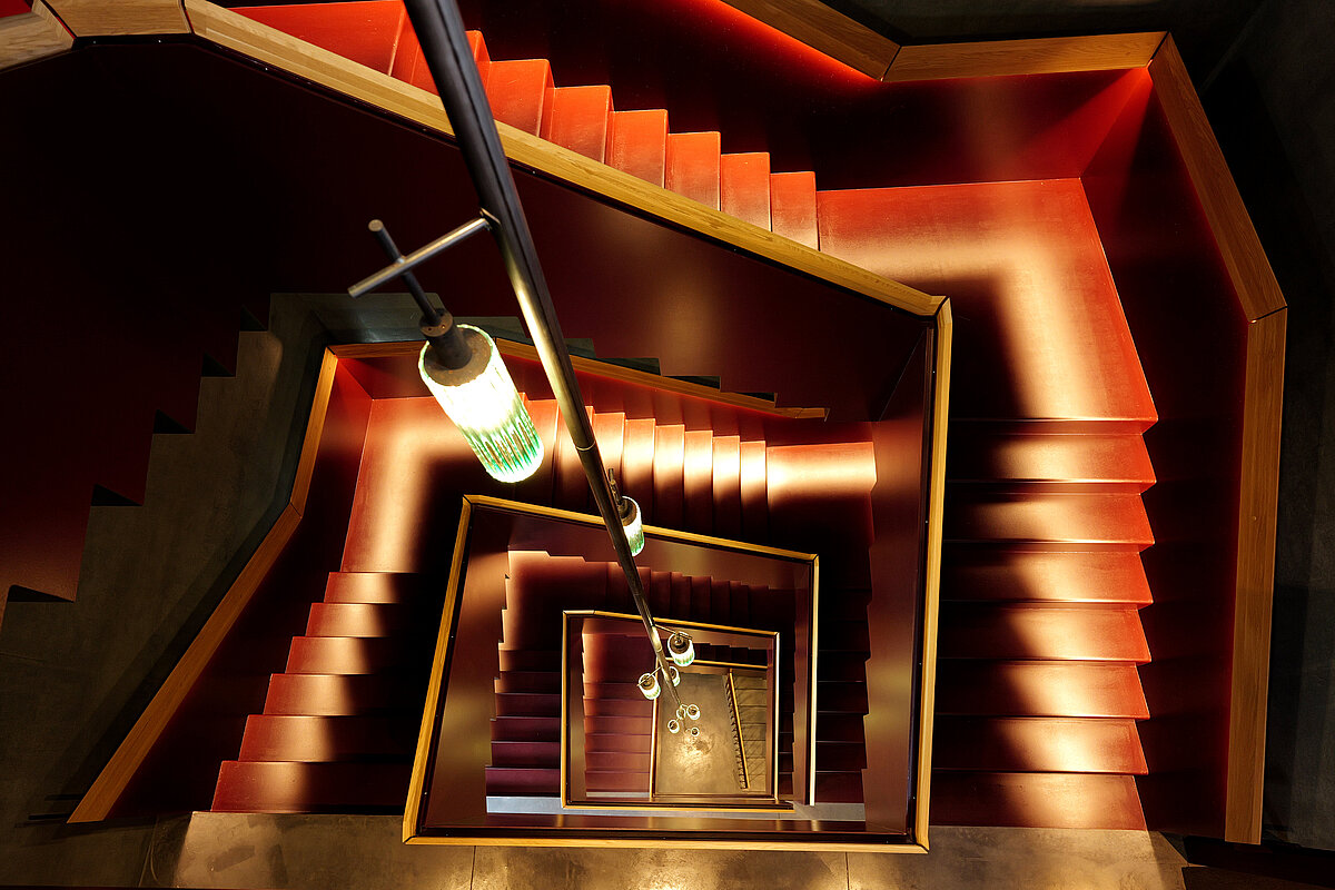 Ansicht eines spiralförmigen Treppenhauses von oben. Die Stufen sind in kräftigem Rot gehalten, während warme Beleuchtung entlang der Treppe für einen dramatischen Effekt sorgt. Ein handlaufähnliches Gestänge verläuft entlang der Mitte.