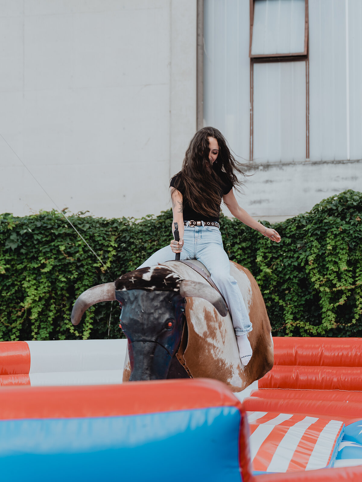 Eine Frau reitet auf einem mechanischen Bullen in einem aufblasbaren Bereich im Freien. Sie trägt Jeans und ein schwarzes Oberteil. Im Hintergrund sind eine grüne Hecke und ein Gebäude zu sehen.