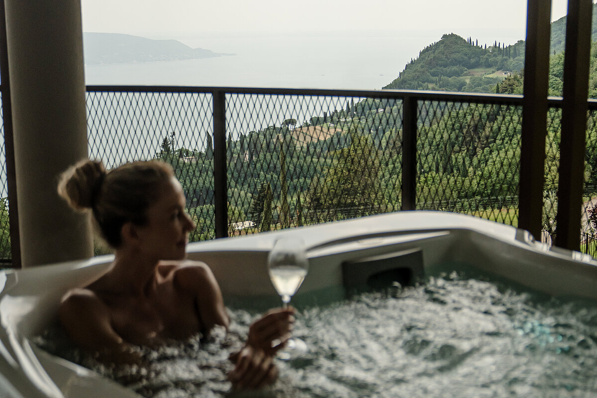 Eine Person entspannt sich mit einem Glas Wein in einem Whirlpool auf einem Balkon. Im Hintergrund ist eine malerische Aussicht auf das Meer und grüne Hügel zu sehen.
