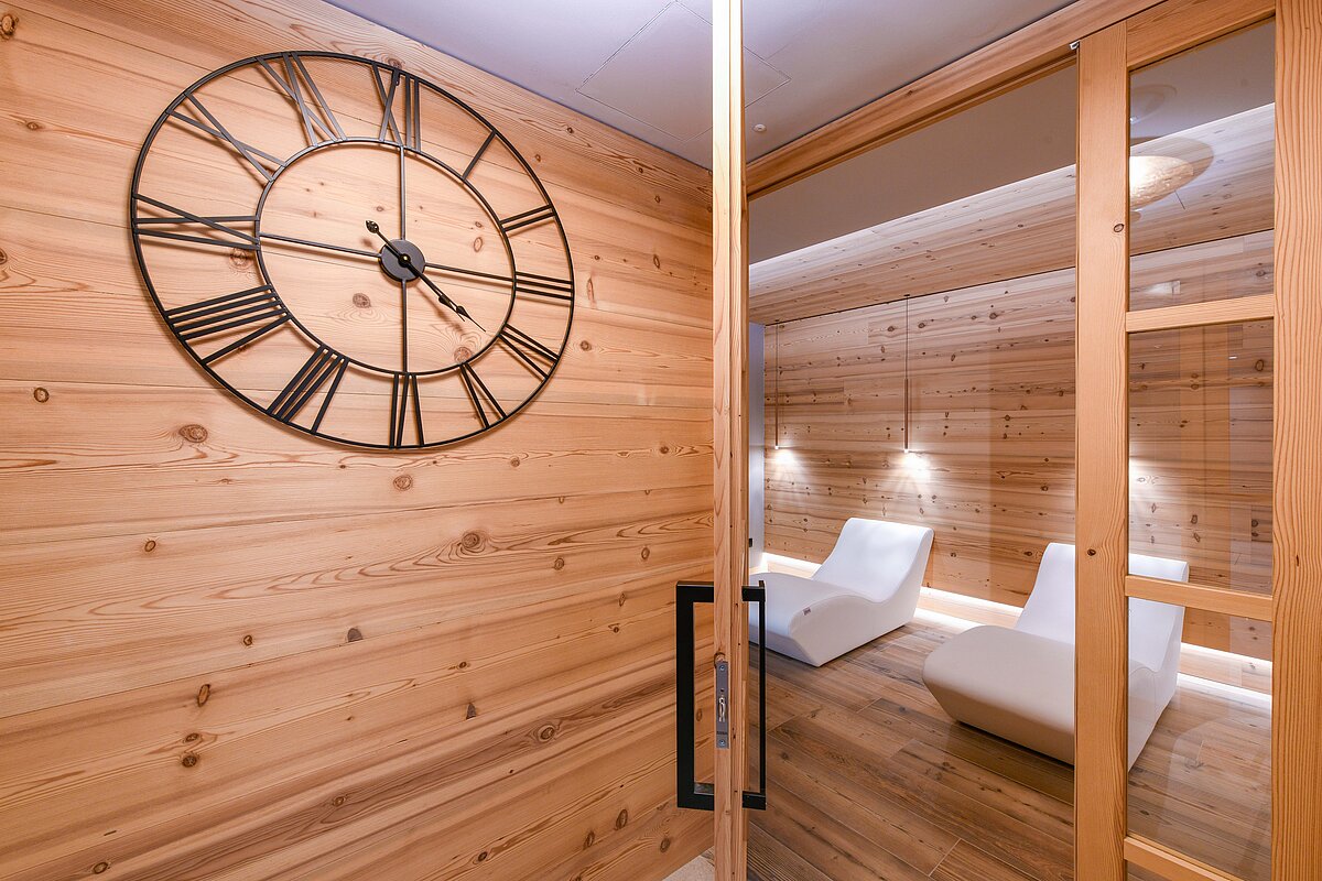 Ein moderner Ruheraum mit Holzwänden und zwei weißen Lounge-Stühlen. An der rechten Wand hängt eine große Wanduhr mit römischen Ziffern. Der Raum hat eine warme Holzoptik und eine ruhige Atmosphäre.