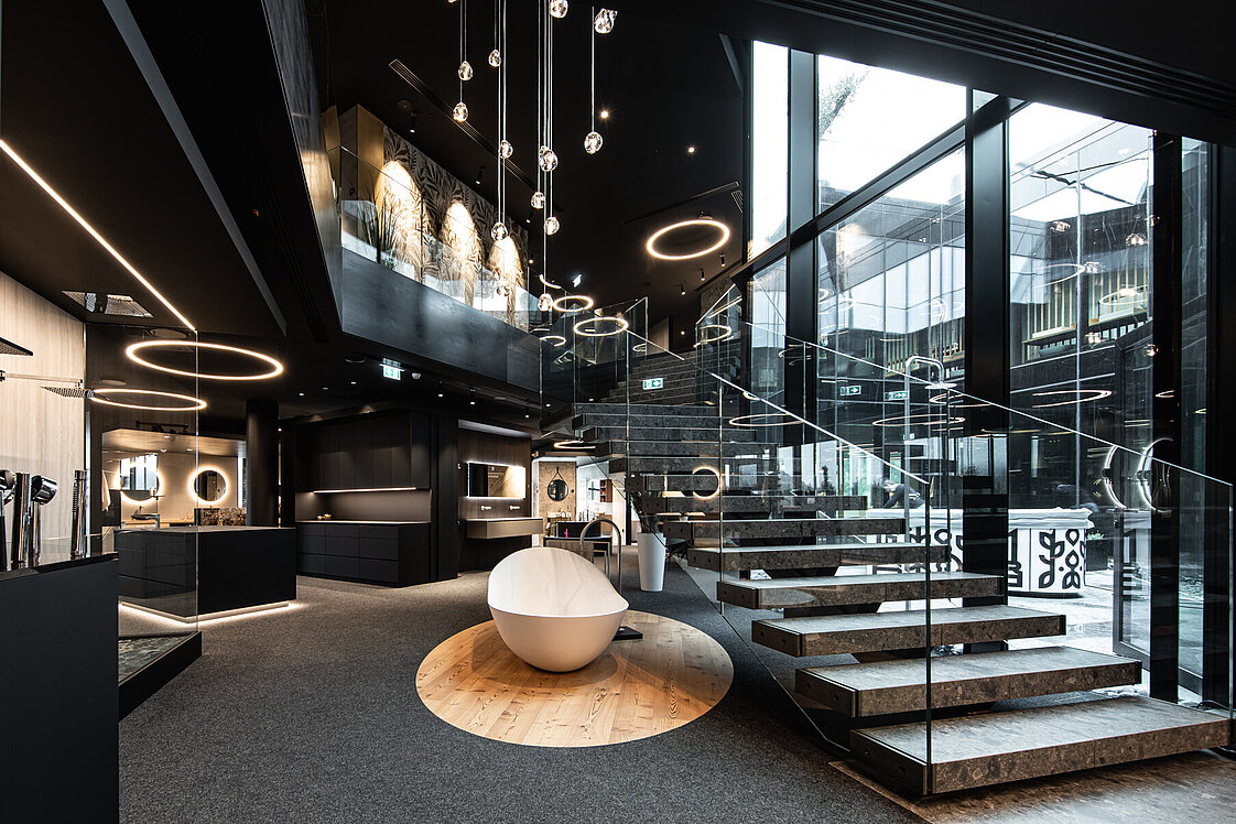 Modernes, stilvolles Innendesign eines Showrooms oder Geschäftsraums mit schwarzen Wänden, großen Glasfronten und kreisförmigen Deckenleuchten. Eine weiße, skulpturale Sitzgelegenheit steht im Vordergrund, während breite Treppen rechts nach oben führen. Luxuriös und offen gestaltet.