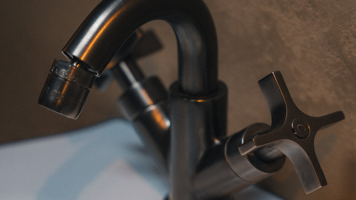 Schwarzer Wasserhahn mit kreuzförmigen Griffen auf einem weißen Waschbecken montiert, vor einer grauen Wand.