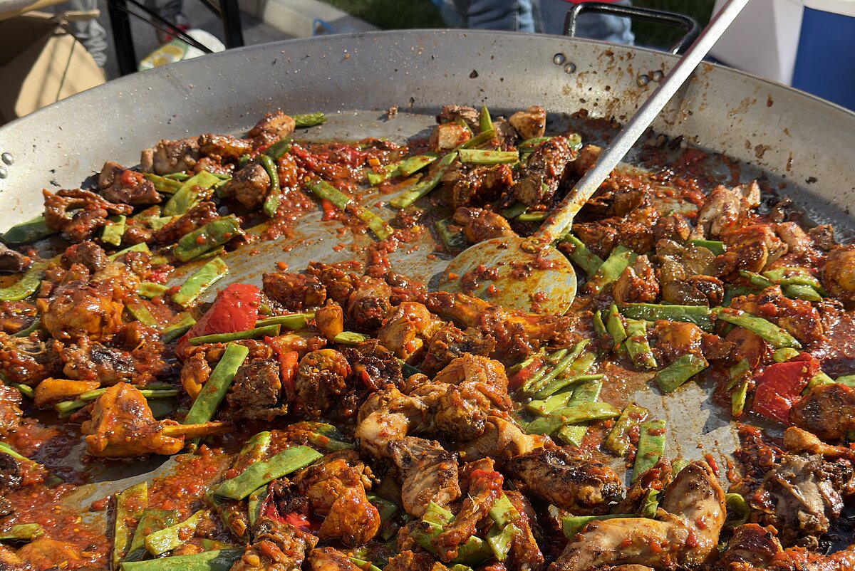 Große Pfanne mit gebratenem Hühnchen, Paprika und grünem Gemüse, auf einer Grillplatte zubereitet. Im Hintergrund sind Personen in roten Schürzen sichtbar.