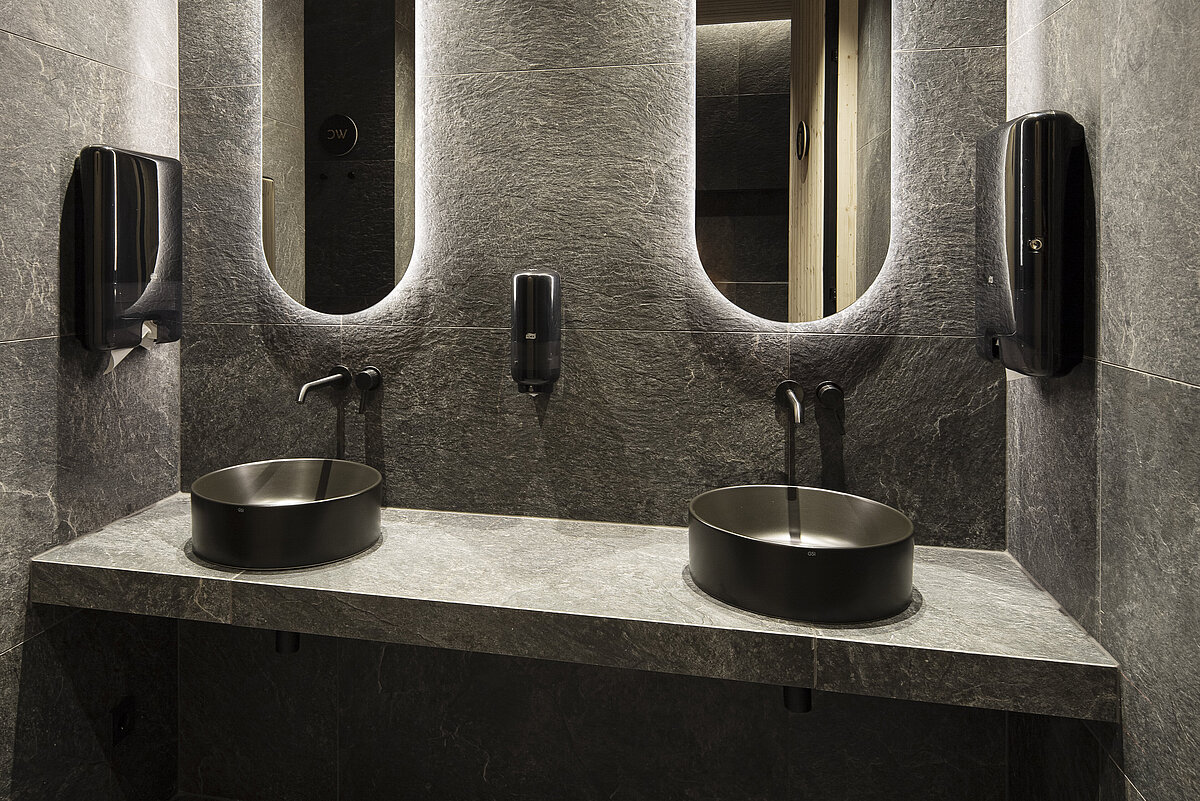 Ein modernes Badezimmer mit zwei runden Waschbecken und schwarzen Armaturen auf einer grauen Steinplatte. Hinter den Becken sind hohe, beleuchtete Spiegel in die Wand eingelassen, die eine elegante Atmosphäre schaffen. Wände und Boden sind einheitlich in dunklen Grautönen gestaltet.
