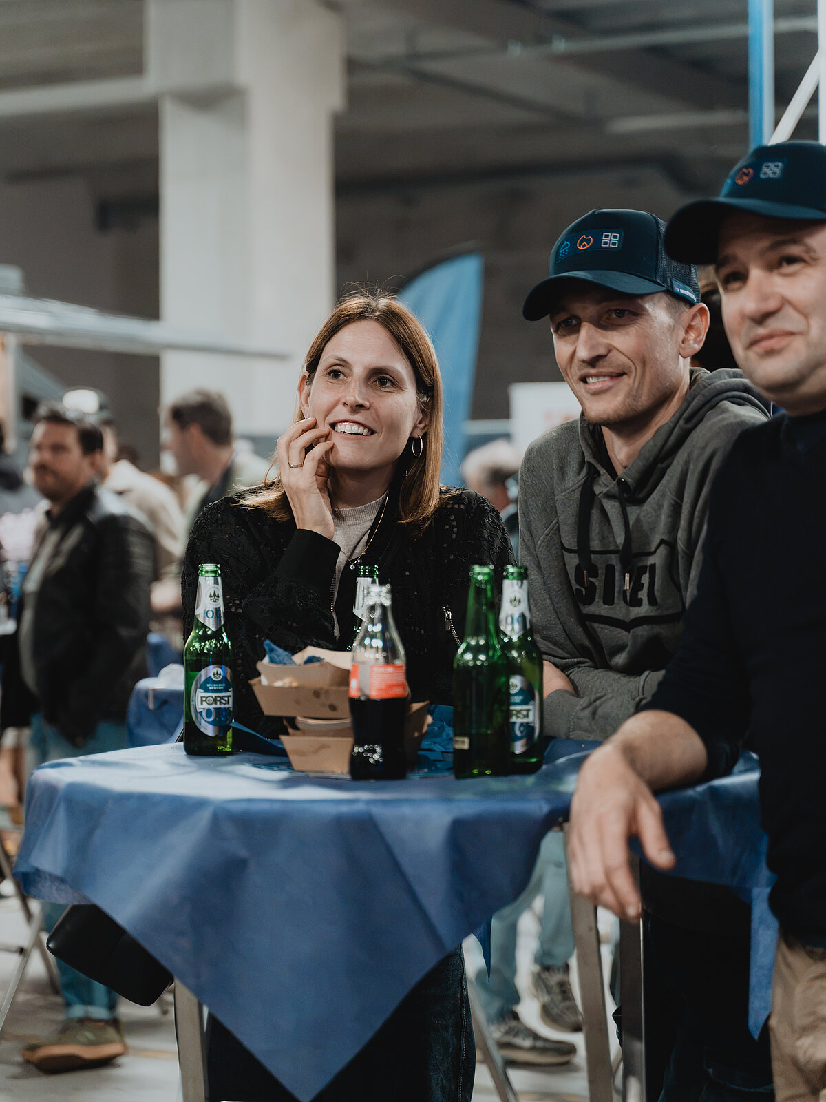 Eine Gruppe von Menschen sitzt lächelnd an einem mit blauer Tischdecke bedeckten Tisch. Auf dem Tisch stehen Bierflaschen und ein Getränk in einer Dose. Im Hintergrund sind weitere Personen und eine industrielle Umgebung zu erkennen.